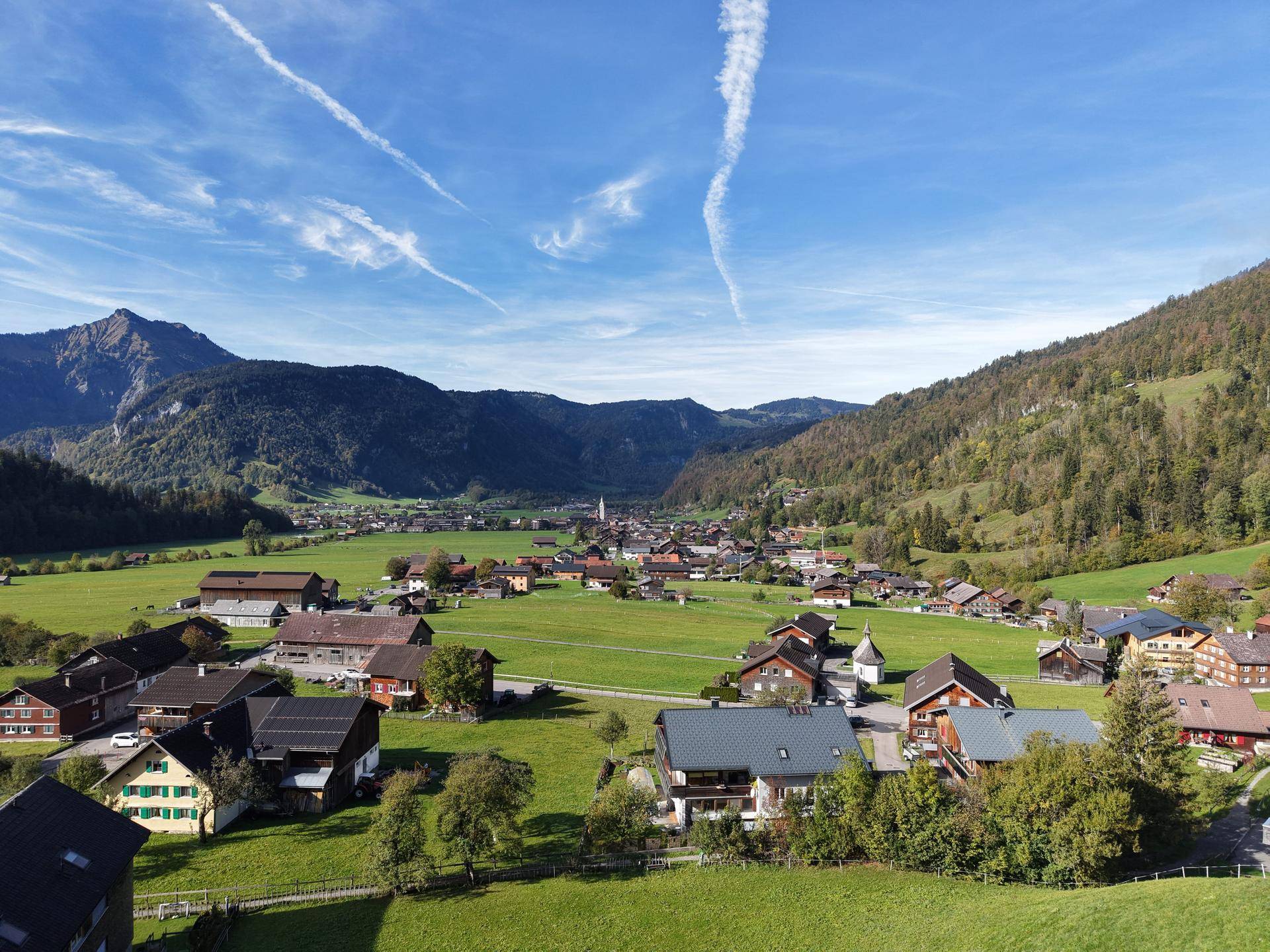 Panoramablick auf ein malerisches Dorf, eingebettet in eine grüne Berglandschaft unter blauem Himmel.