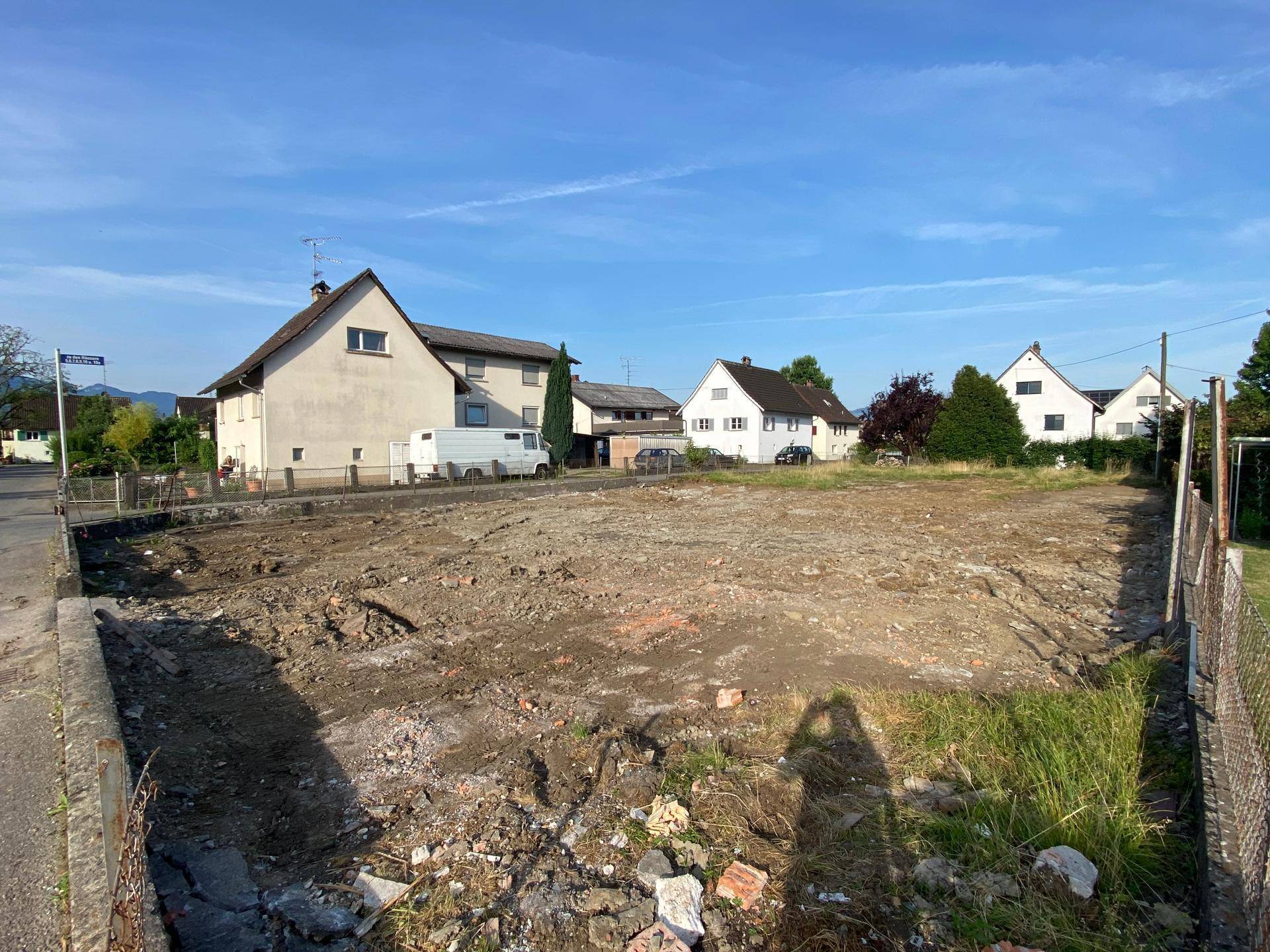 Geräumiges Baugrundstück an einer Straße, mit Häusern und Bergen im Hintergrund unter blauem Himmel.
