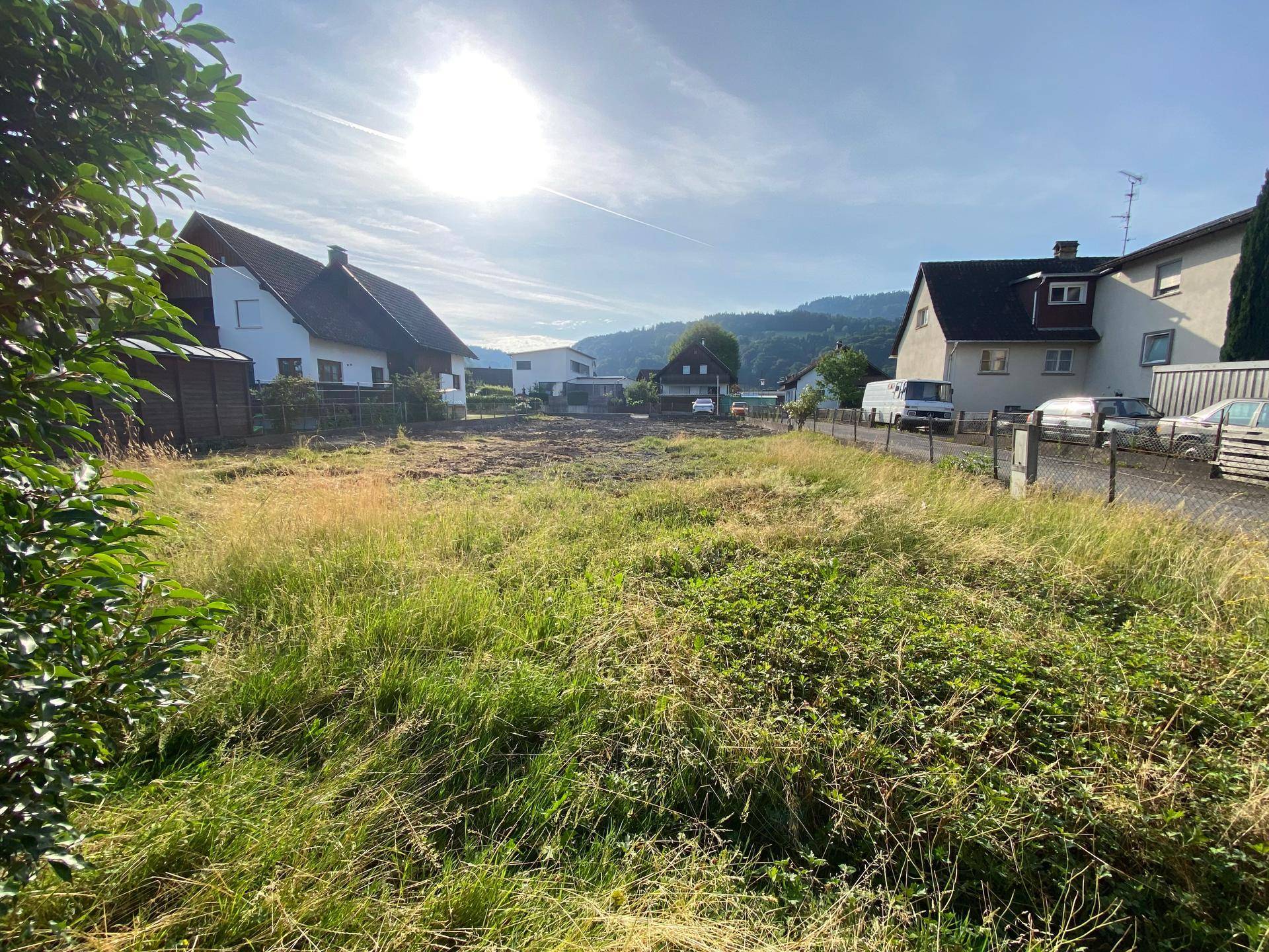Grünes Baugrundstück mit hohem Gras, umgeben von Wohnhäusern unter strahlendem Sonnenschein.