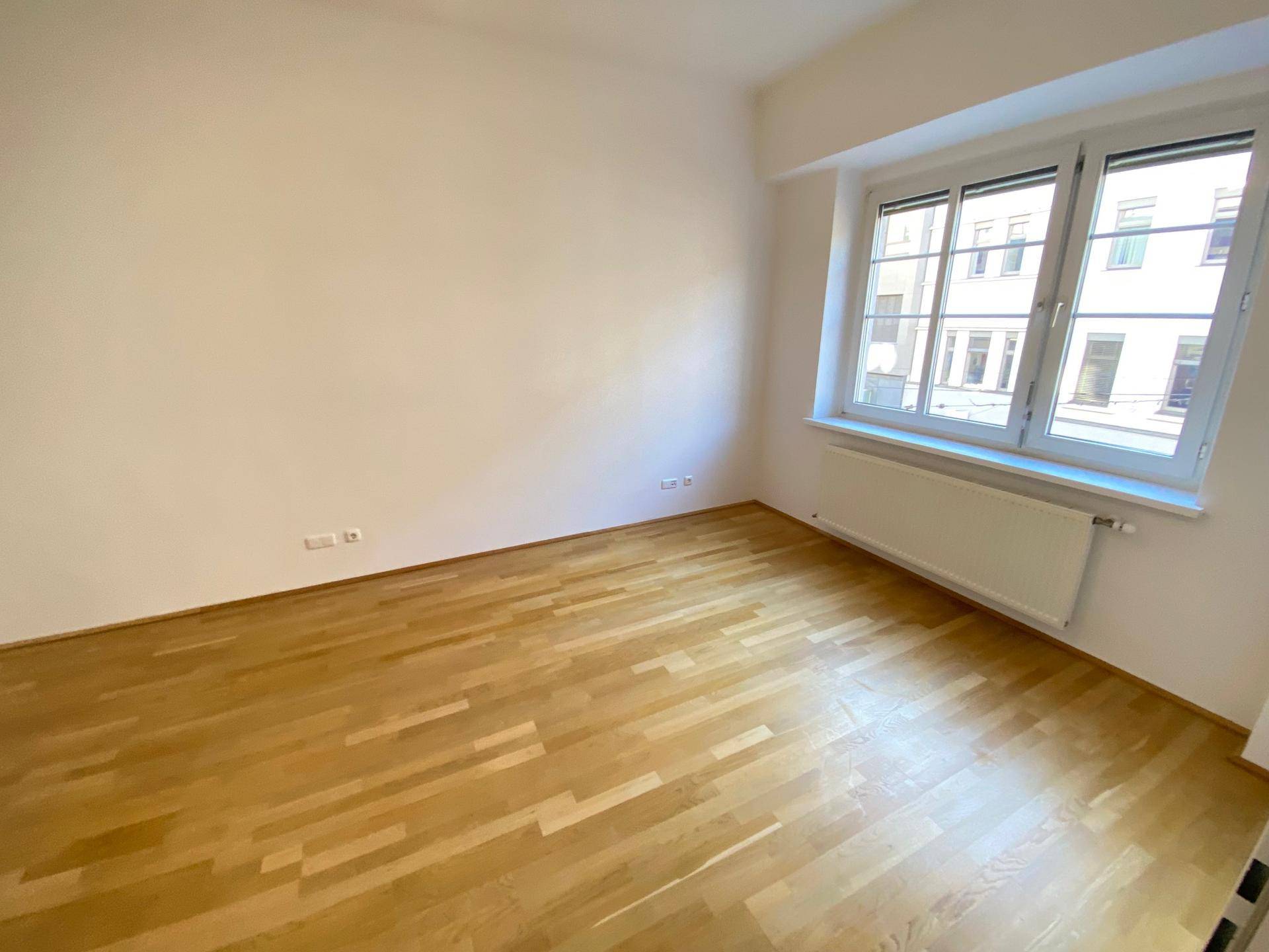 Unmöbliertes Zimmer mit hellem Holzboden und einem großen Fenster, das den Raum mit Tageslicht durchflutet.