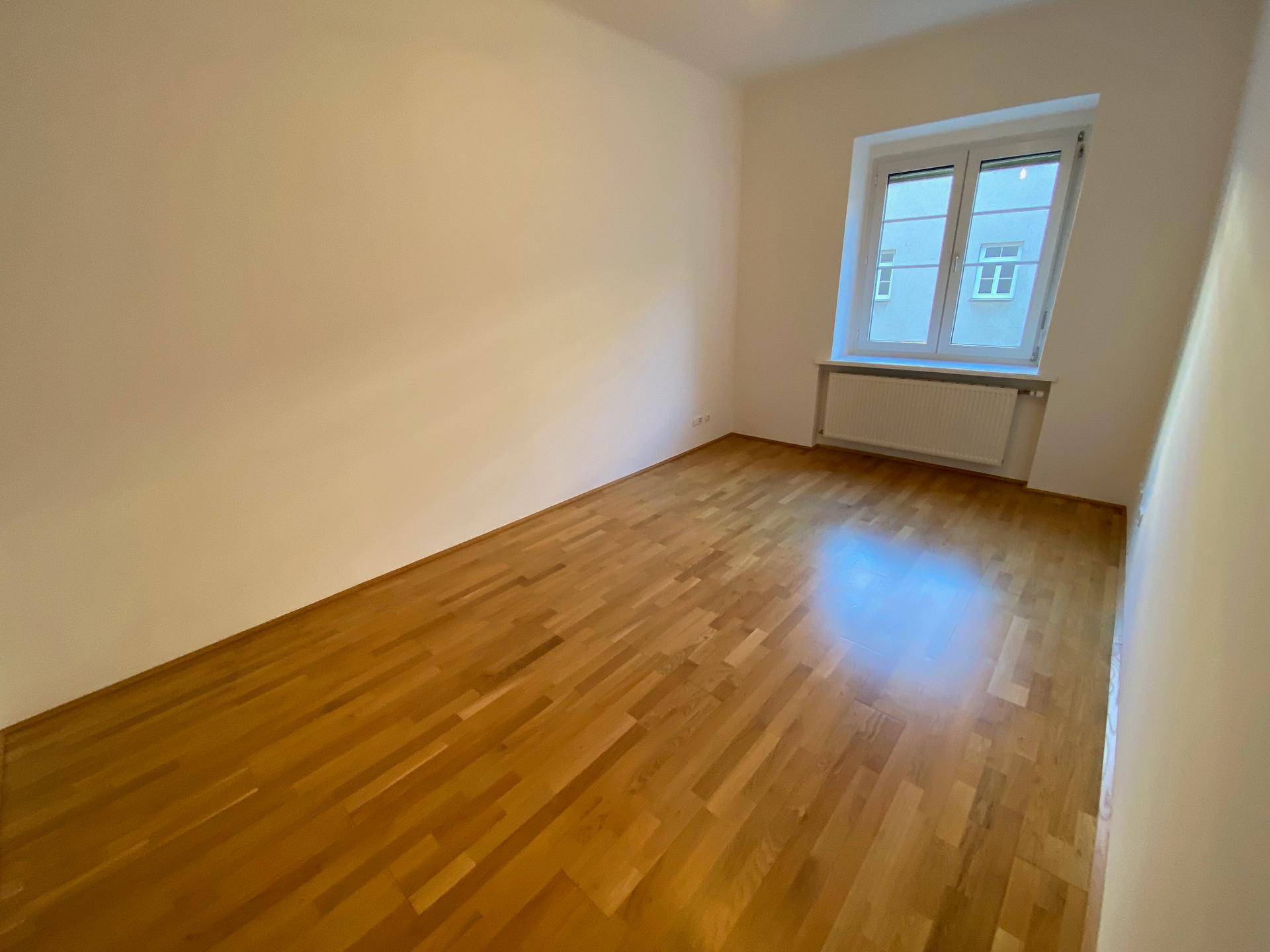 Helles, leeres Zimmer mit warmem Holzboden und einem großen Fenster, das für eine angenehme Atmosphäre sorgt.