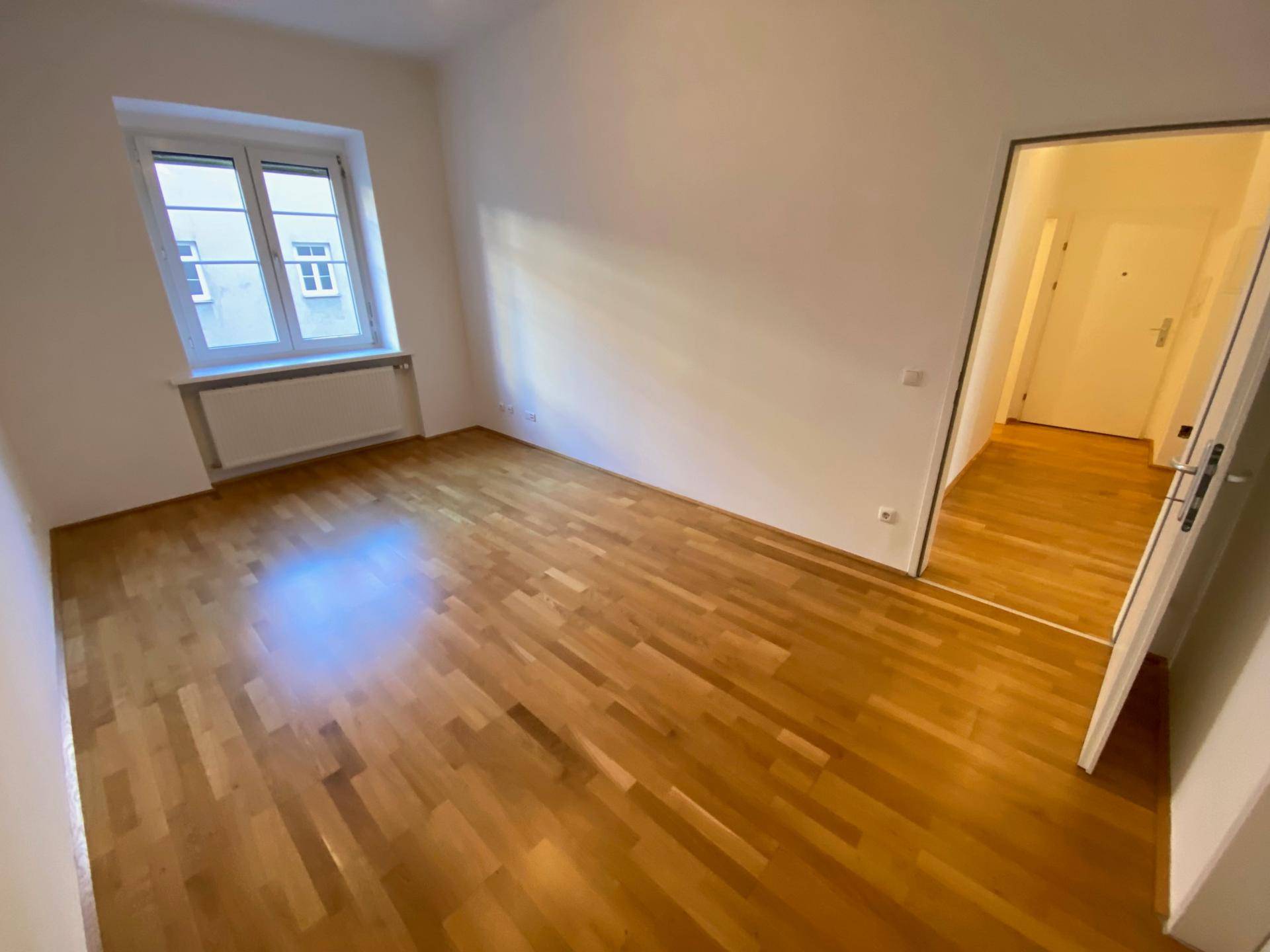 Leeres Zimmer mit Holzboden und einem Fenster, das viel natürliches Licht hereinlässt und einen Blick nach draußen bietet.