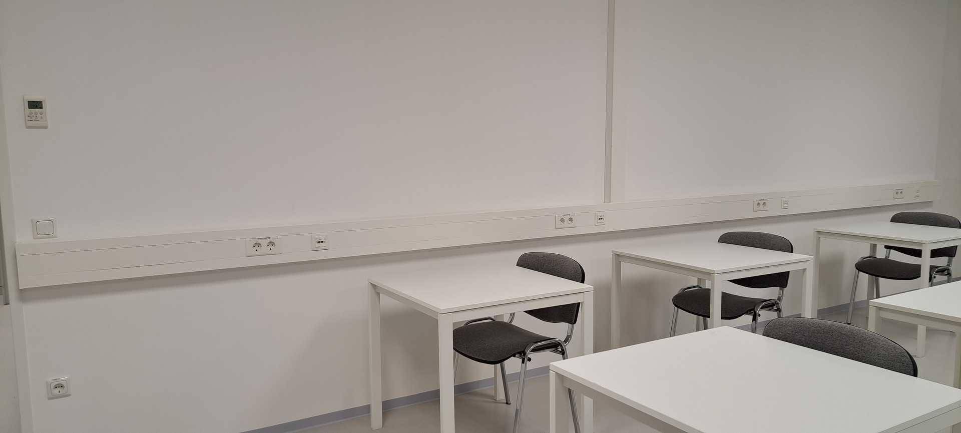 Schulungsraum mit weißen Tischen und Stühlen, ausgestattet mit zahlreichen Steckdosen entlang der Wand.