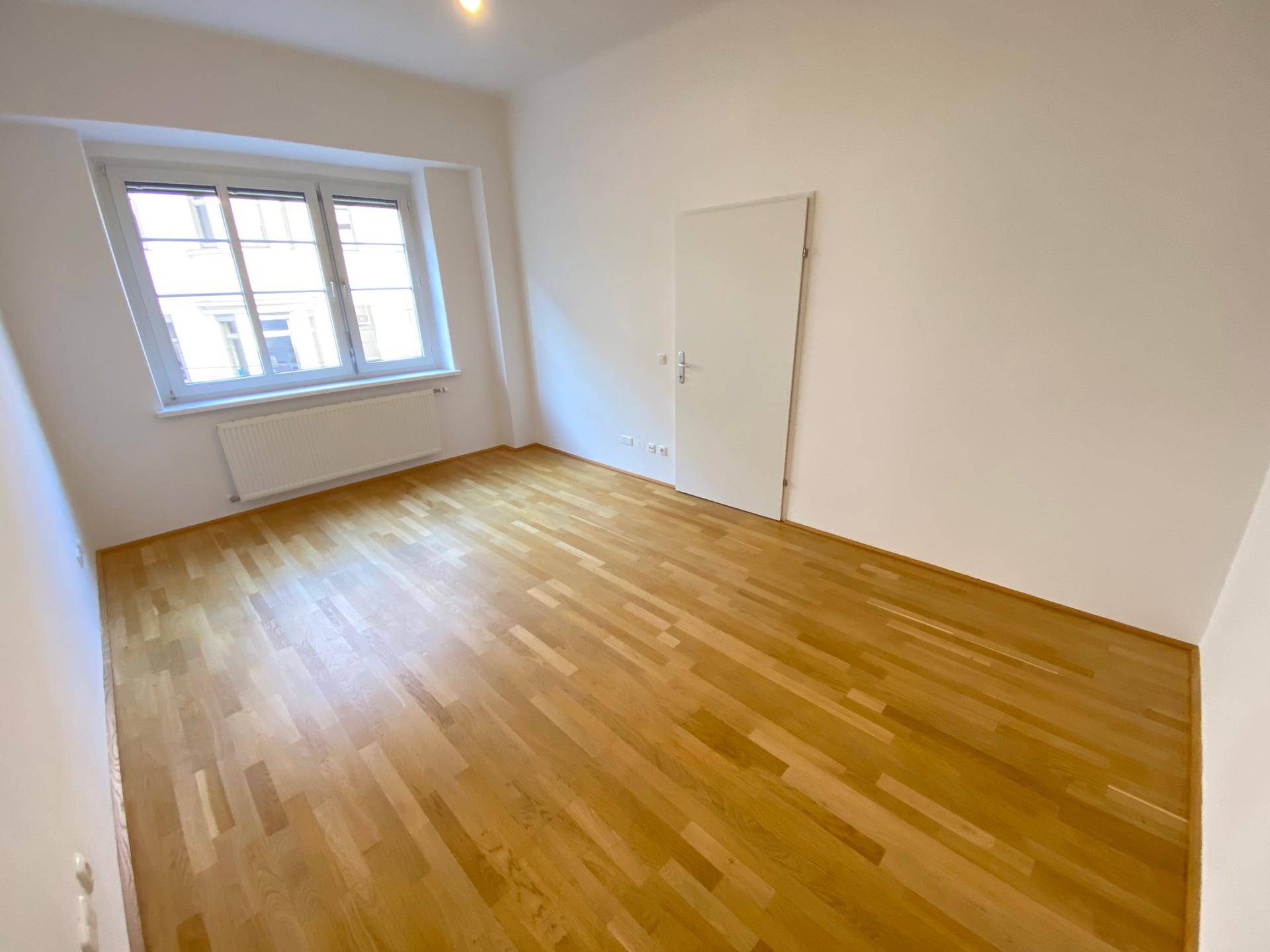 Geräumiges, leeres Zimmer mit hellem Holzboden und einem großen Fenster, das viel Tageslicht bietet.