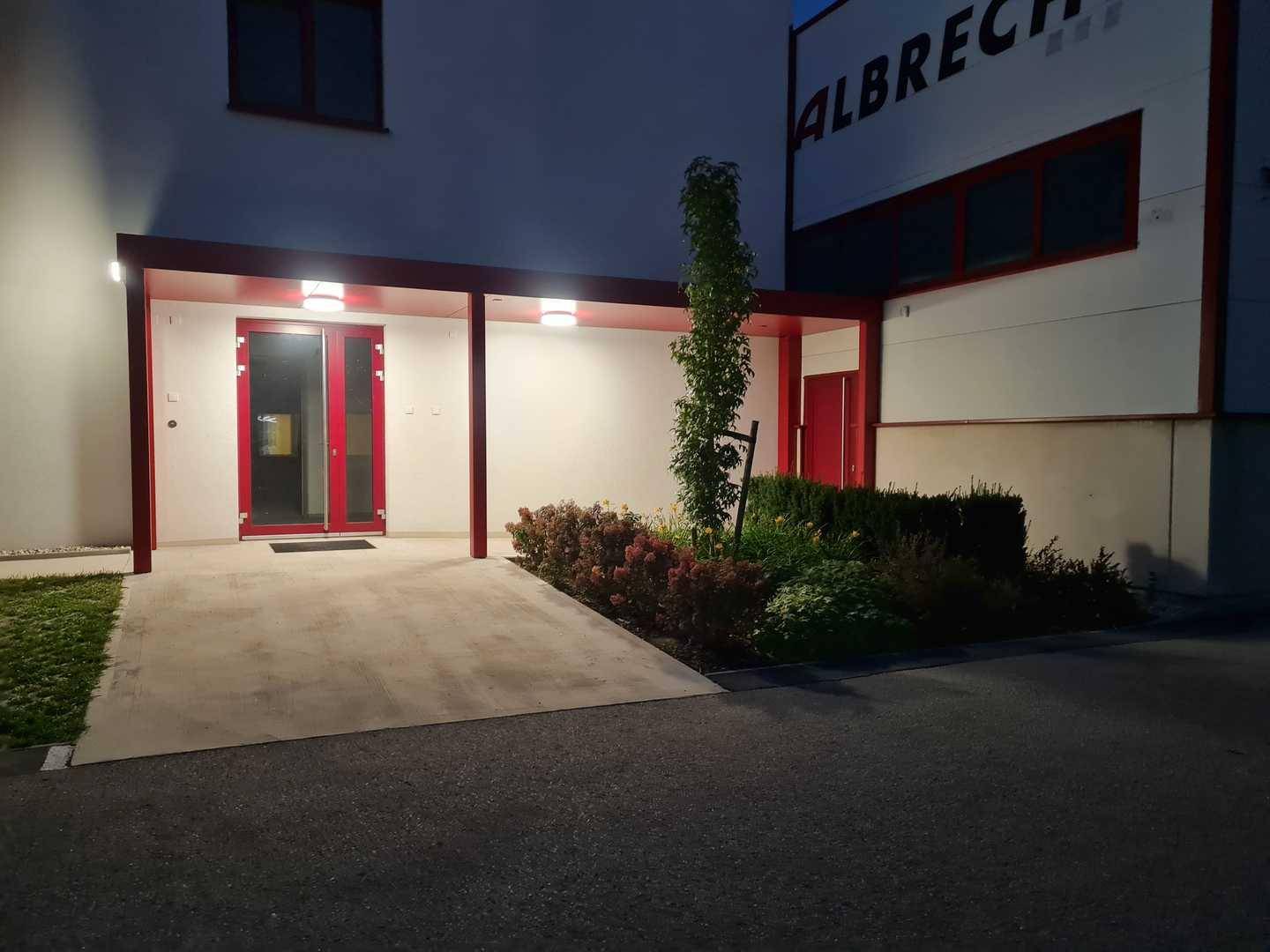Beleuchteter Eingangsbereich eines modernen Gebäudes mit roten Türen und gepflegter Bepflanzung am Abend.