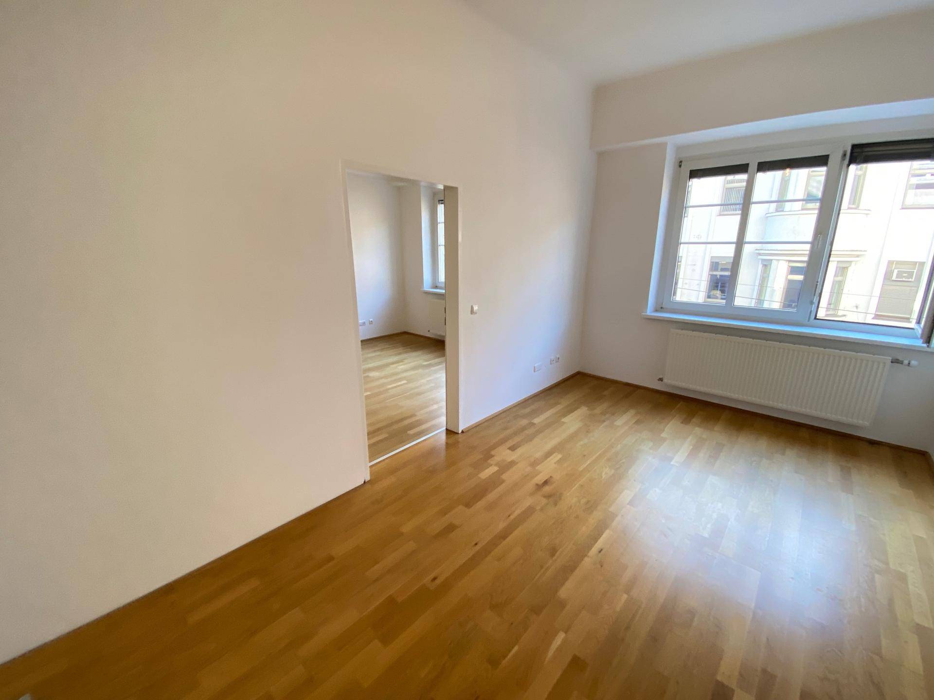 Unmöblierter Wohnbereich mit glänzendem Holzboden und Blick in einen weiteren Raum durch eine offene Tür.