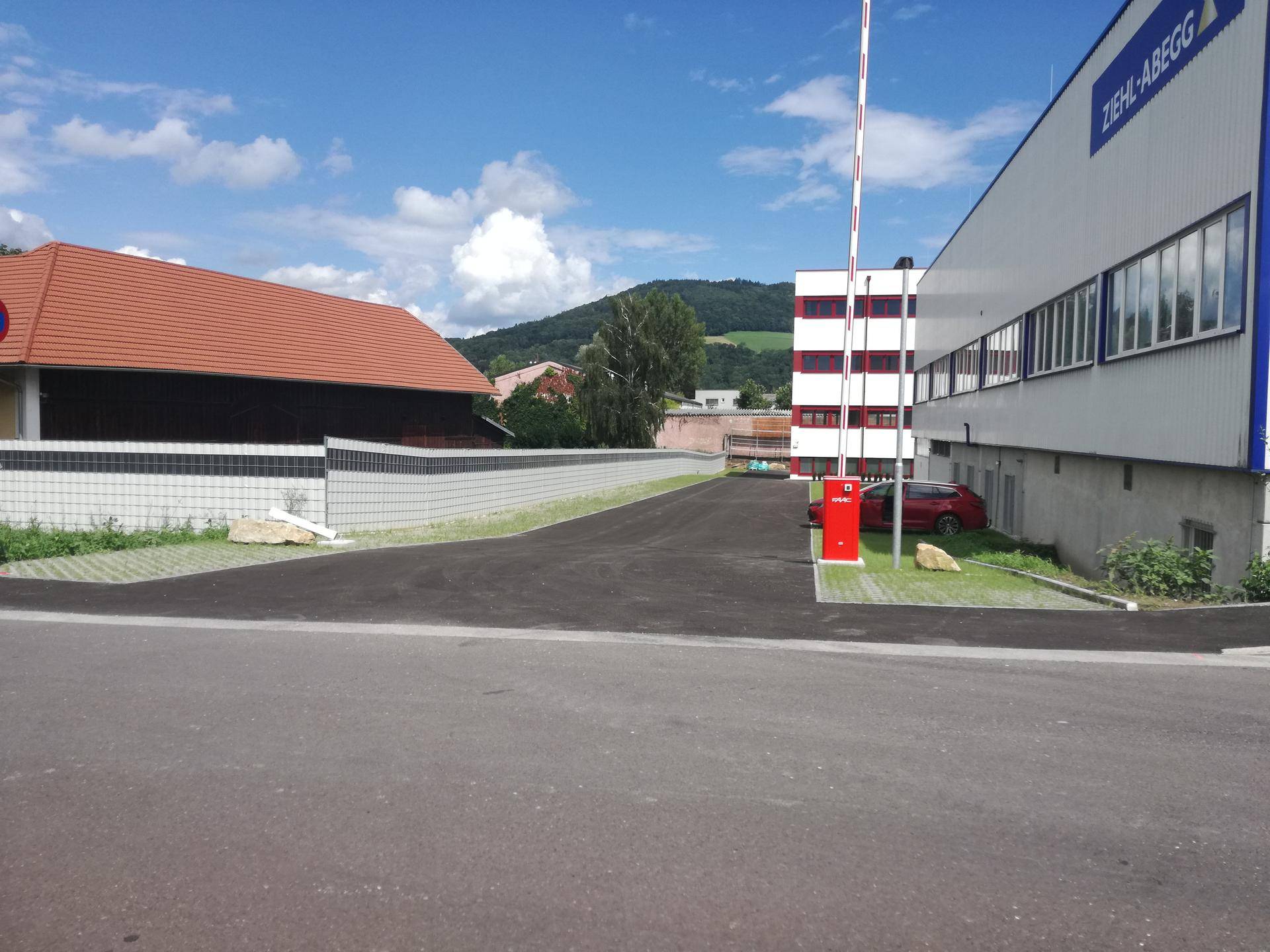 Außenansicht eines Gewerbegebiets mit Zufahrt, Schranke und angrenzenden Gebäuden unter blauem Himmel.