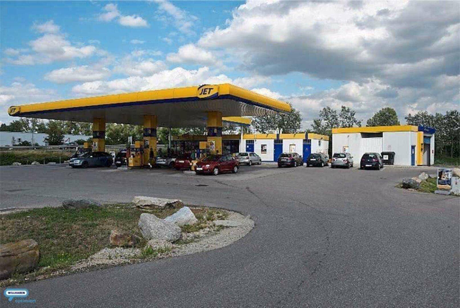 Luftaufnahme einer Tankstelle mit mehreren Fahrzeugen und angrenzenden Servicegebäuden unter einem bewölkten Himmel.