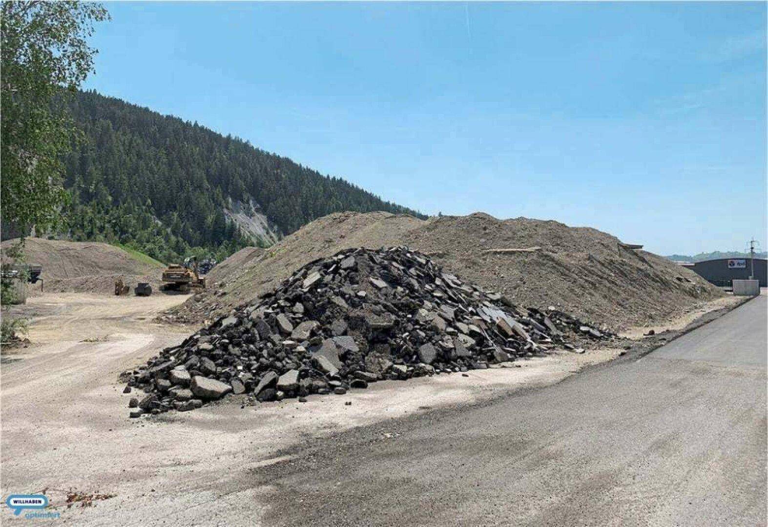 Große Erdhügel und Schutthalden auf einem Gelände mit bewaldeten Bergen unter klarem blauem Himmel.