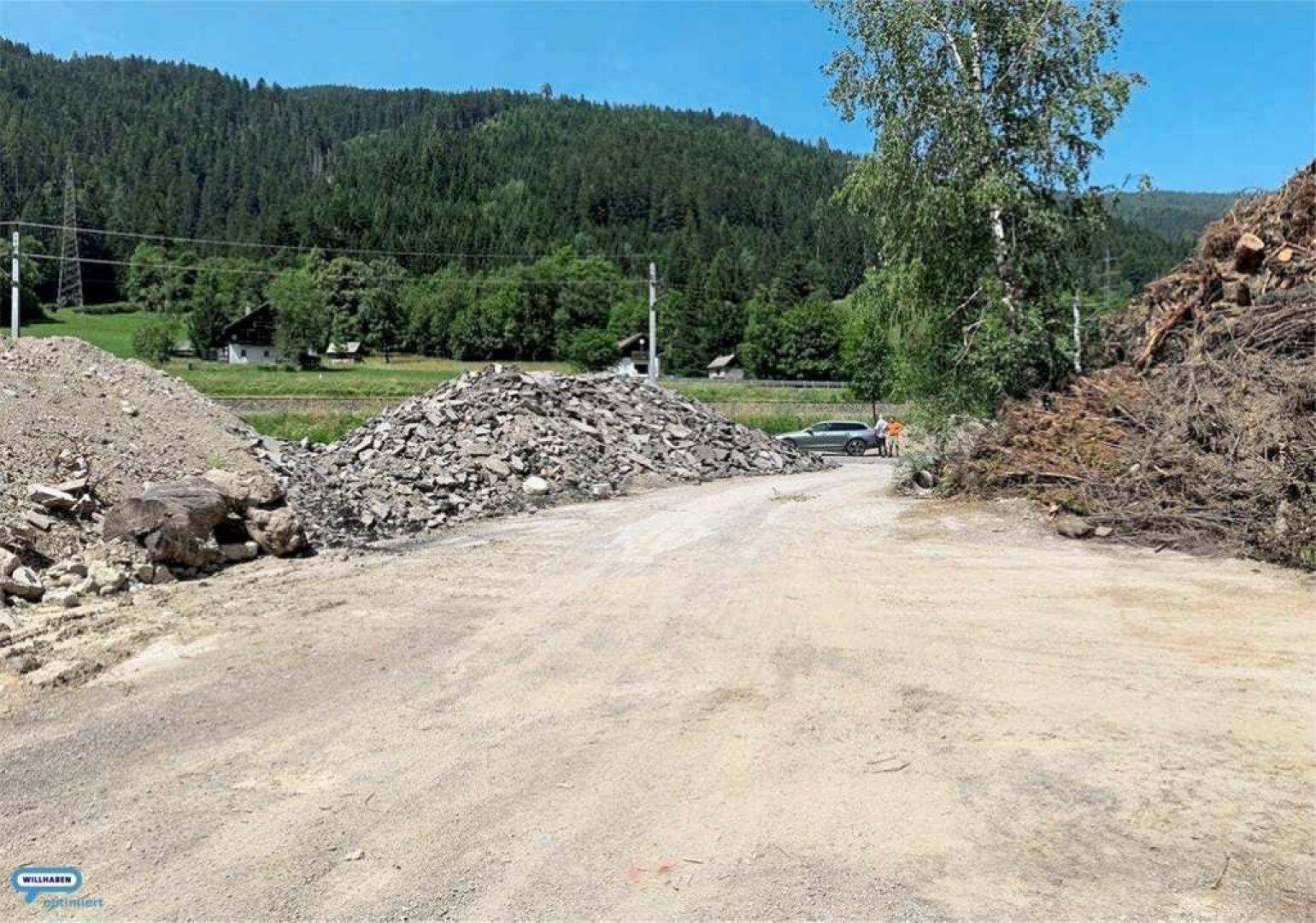 Schotterweg zwischen großen Haufen von Bauschutt und Holzabfällen, umgeben von grünen Wäldern und Bergen.