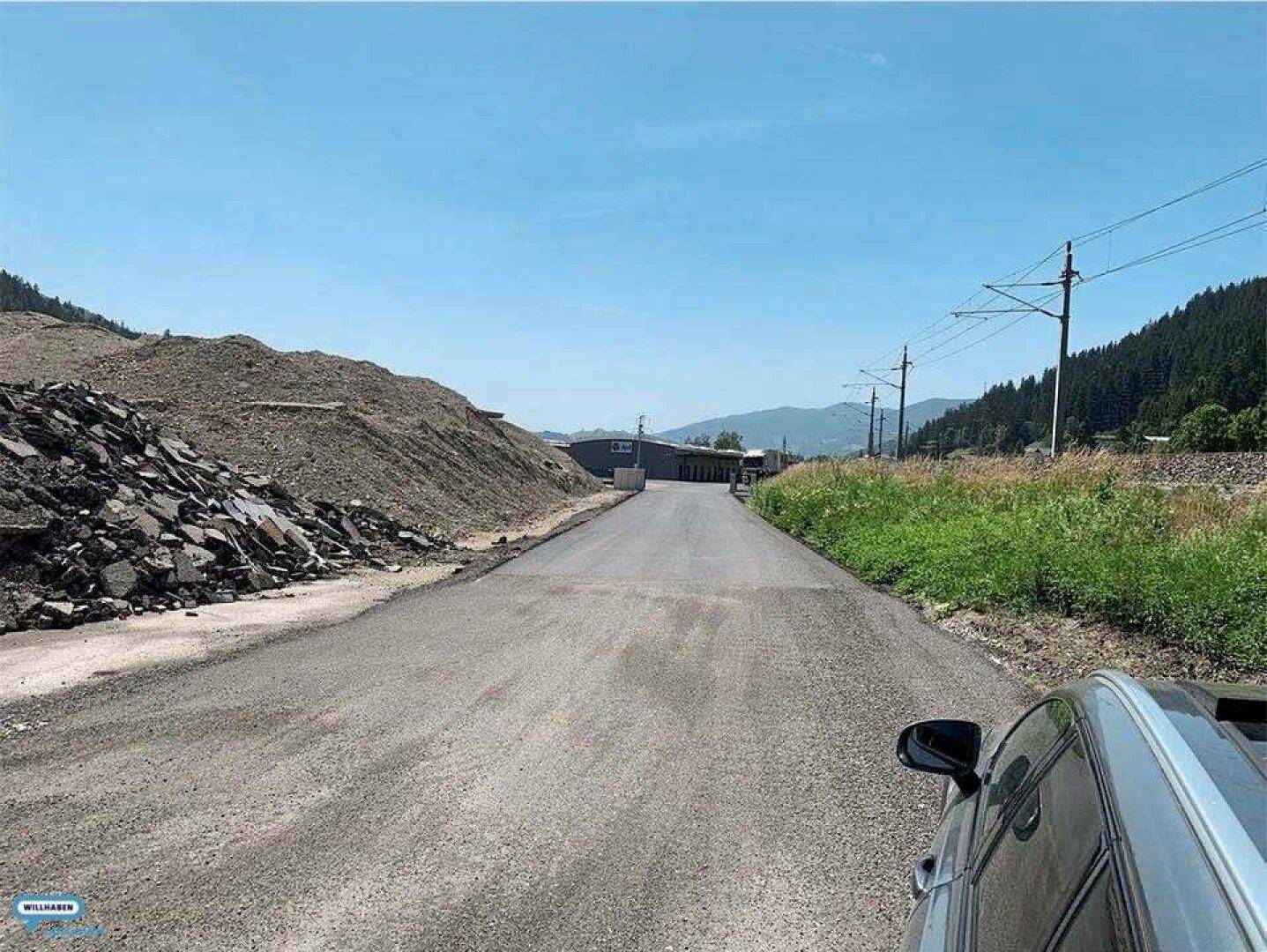 Blick entlang einer asphaltierten Straße mit Erdhügeln, Stromleitungen und Bergen in der Ferne.