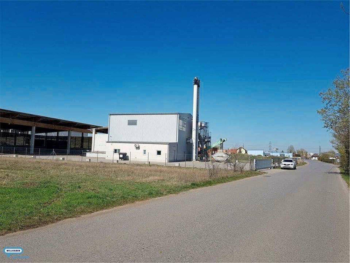 Industriegebäude mit hohem Schornstein neben einer Straße und einem grünen Feld unter blauem Himmel.