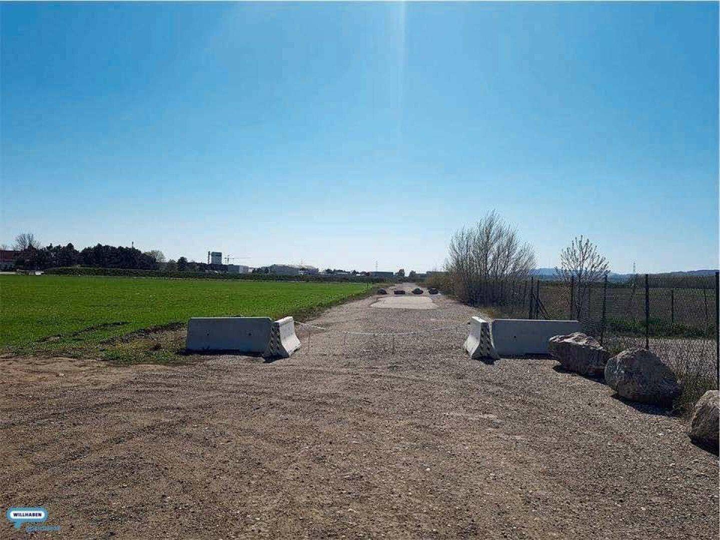 Unbefestigter Weg mit Betonbarrieren, der zu einem weiten Feld führt, umgeben von Natur.
