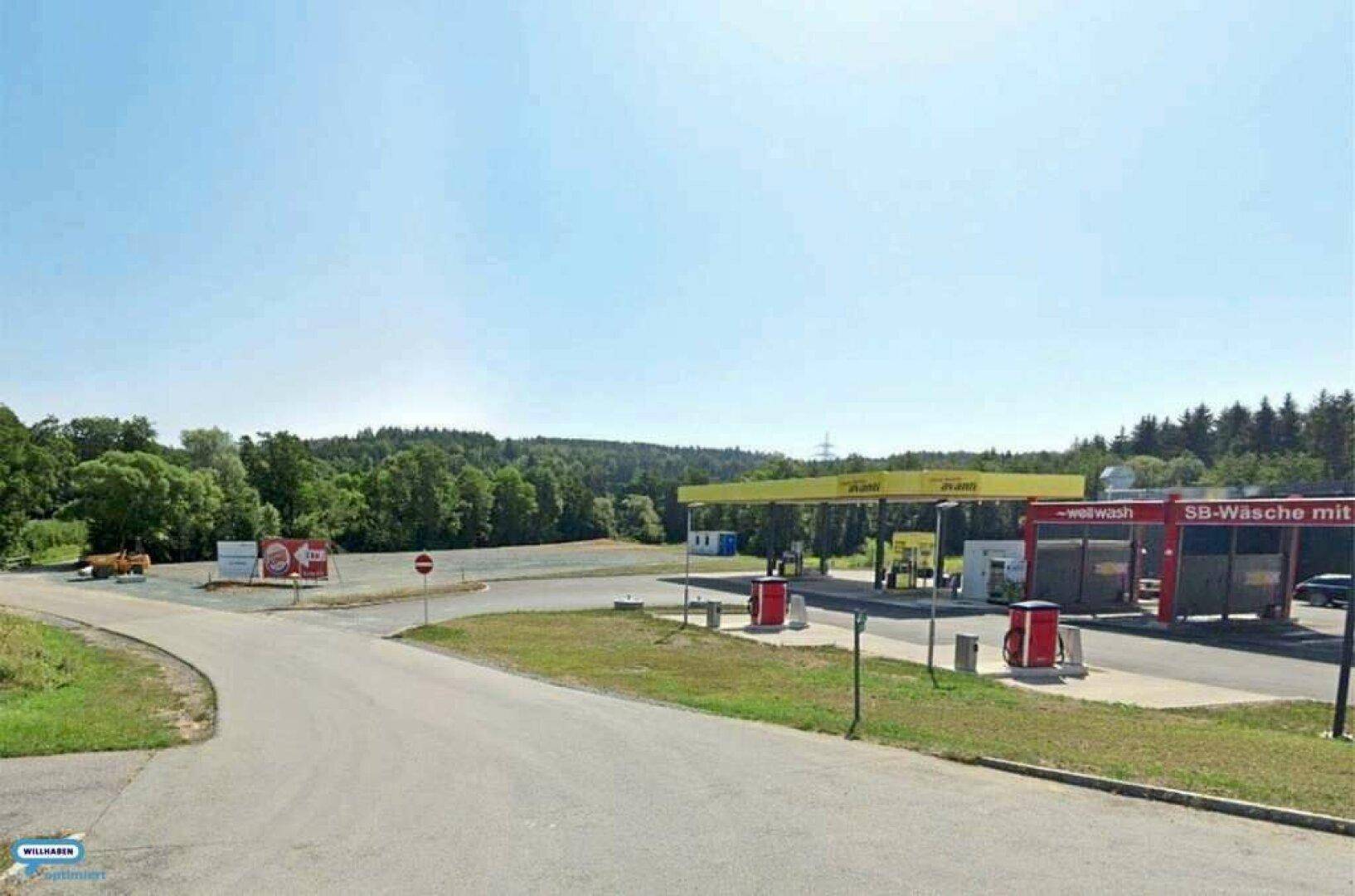 Weitläufiges Gelände mit einer Tankstelle und einer Autowaschanlage, umgeben von Bäumen und Natur unter blauem Himmel.