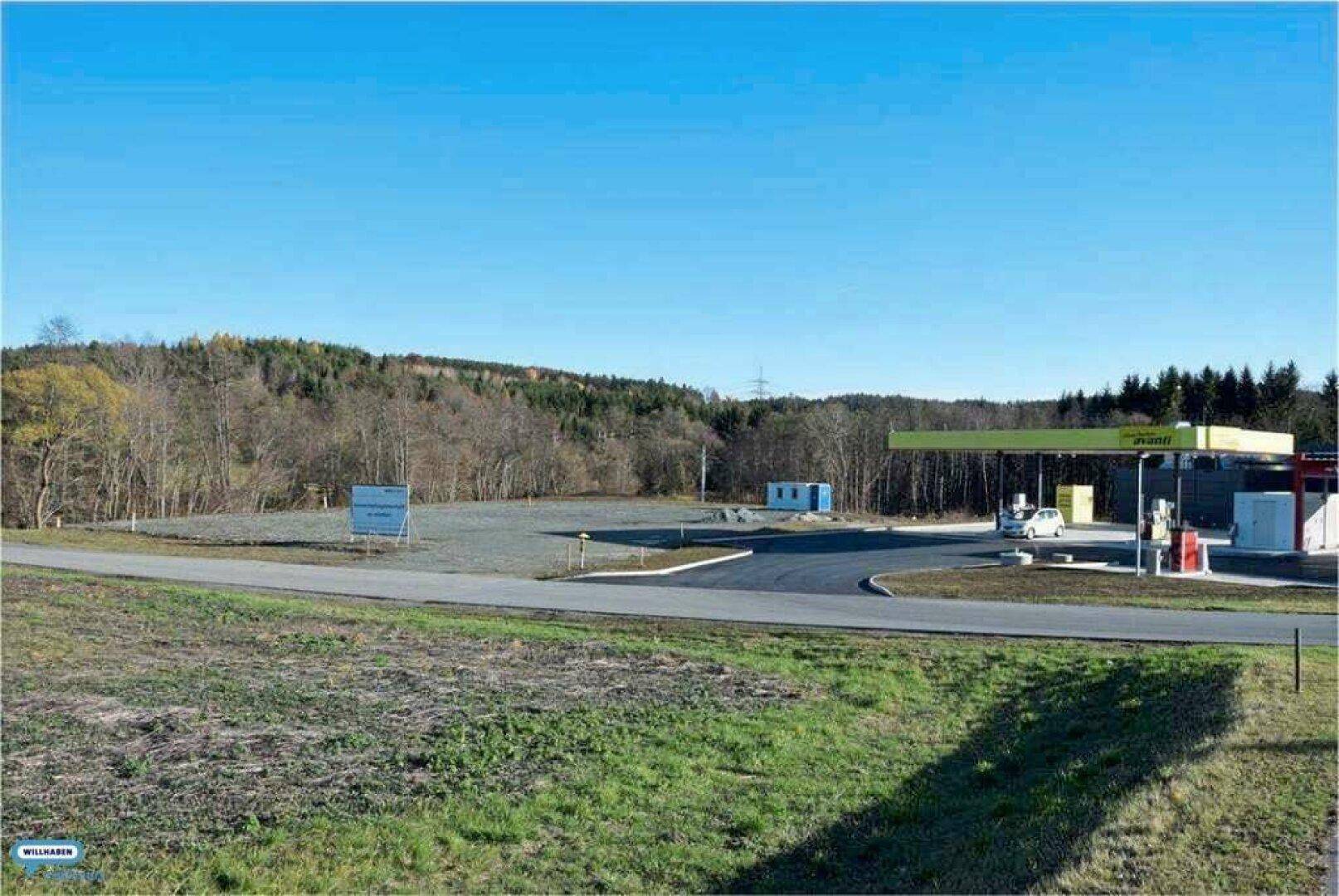 Eine weite, ungenutzte Fläche neben einer Tankstelle, umgeben von Wald und ländlicher Landschaft unter klarem Himmel.
