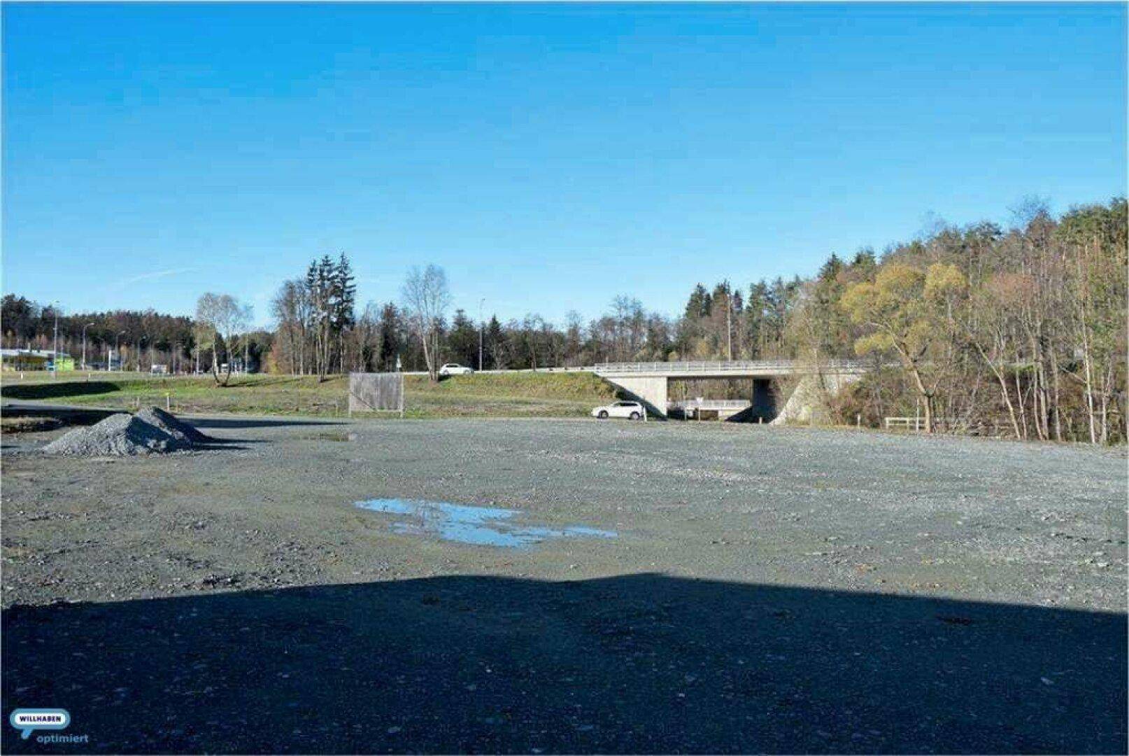 Eine große, unbefestigte Fläche mit einer Pfütze und einer Brücke im Hintergrund, umgeben von Bäumen.