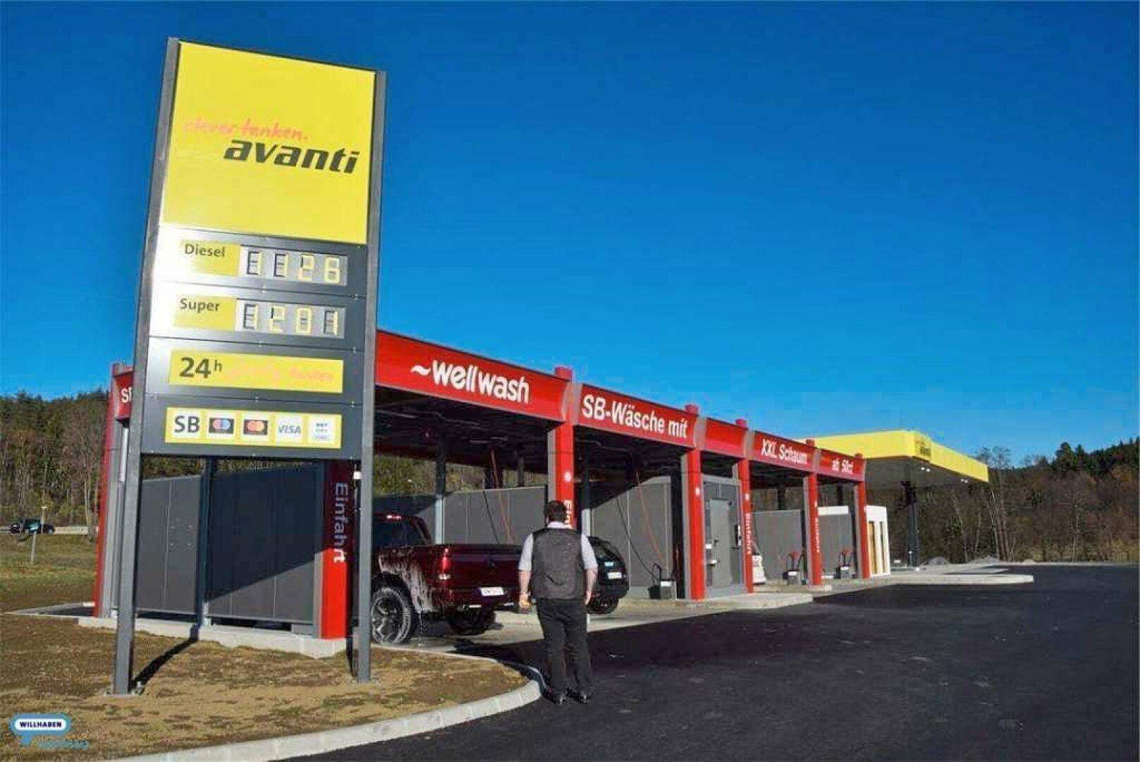 Eine moderne Tankstelle mit Preisanzeige und einer SB-Waschanlage unter strahlend blauem Himmel.