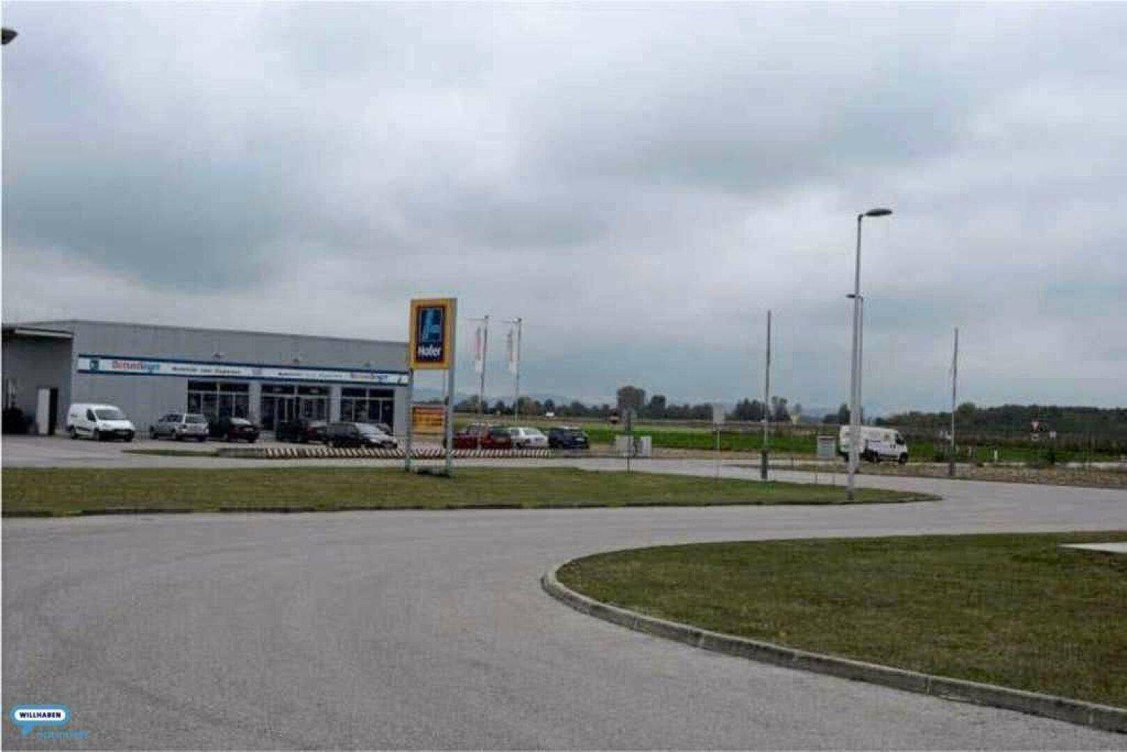 Ansicht eines Hofer-Supermarktes mit Parkplatz und Zufahrtsstraße unter einem bewölkten Himmel.
