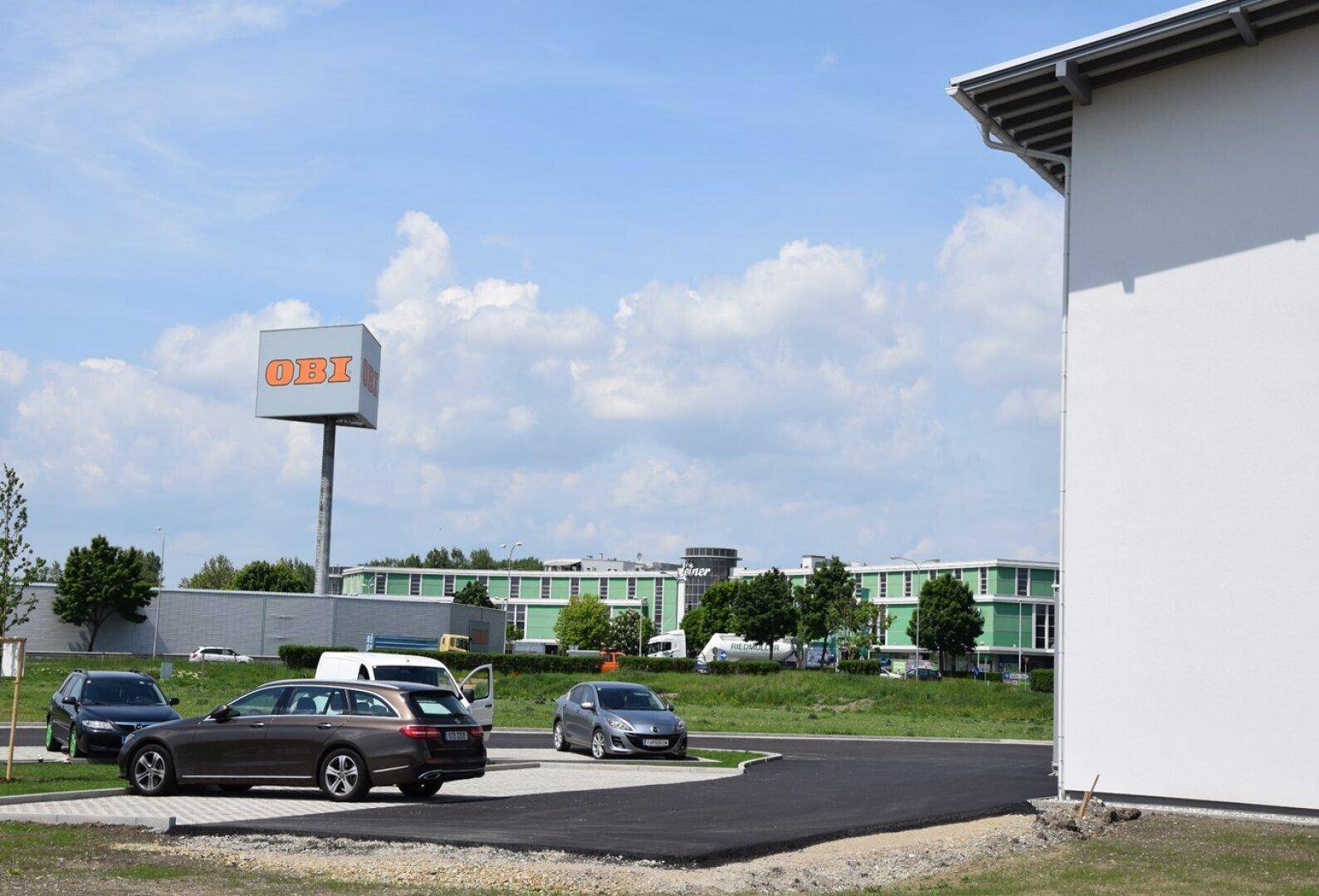 Parkplatzbereich vor einem OBI-Baumarkt mit weiteren Geschäftsgebäuden im Hintergrund unter einem klaren blauen Himmel.
