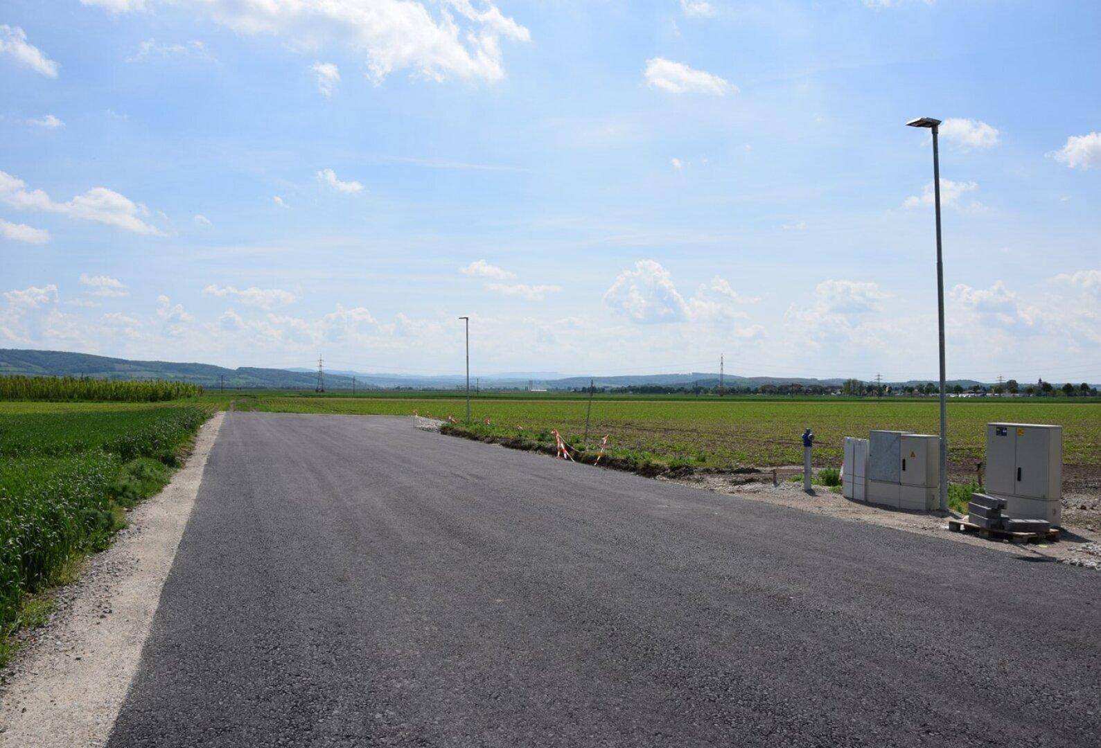 Eine neu asphaltierte Straße führt durch weite grüne Felder unter einem klaren blauen Himmel mit ländlicher Aussicht.