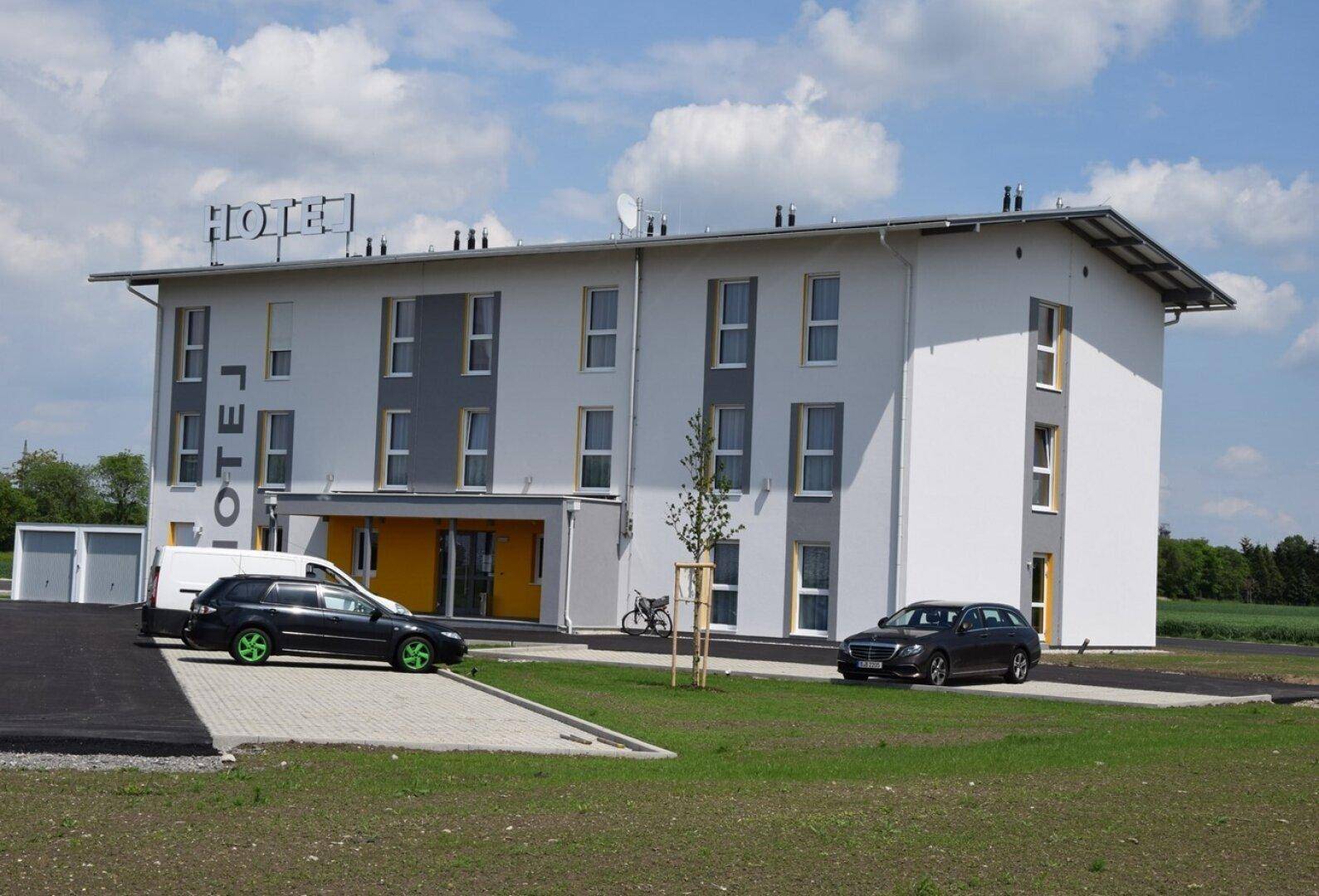Modernes Hotelgebäude mit weißer und grauer Fassade, gelben Akzenten und Parkplätzen davor unter blauem Himmel.