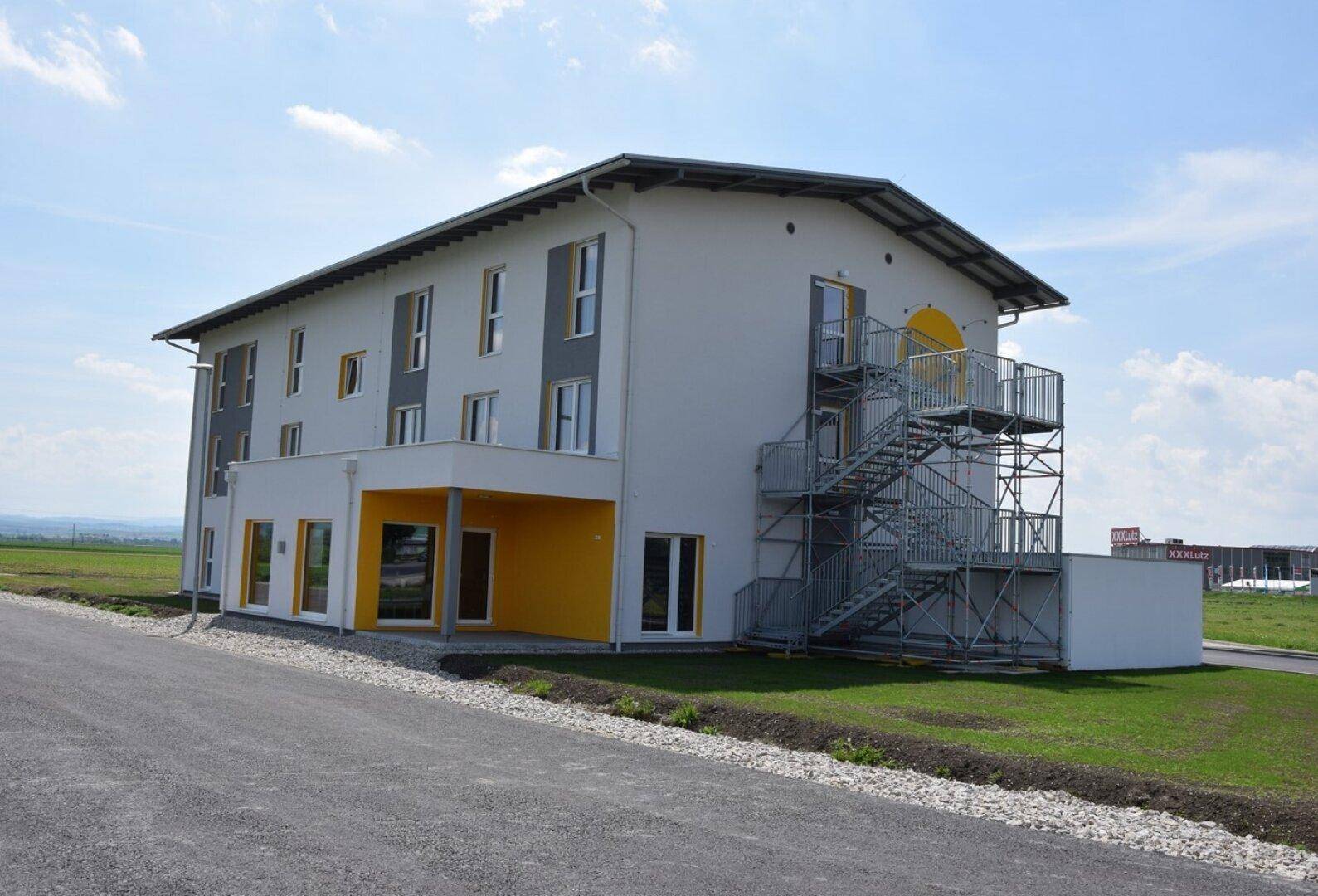 Seitenansicht des modernen Hotelgebäudes mit Außentreppe und gelben Akzenten an der Fassade unter blauem Himmel.