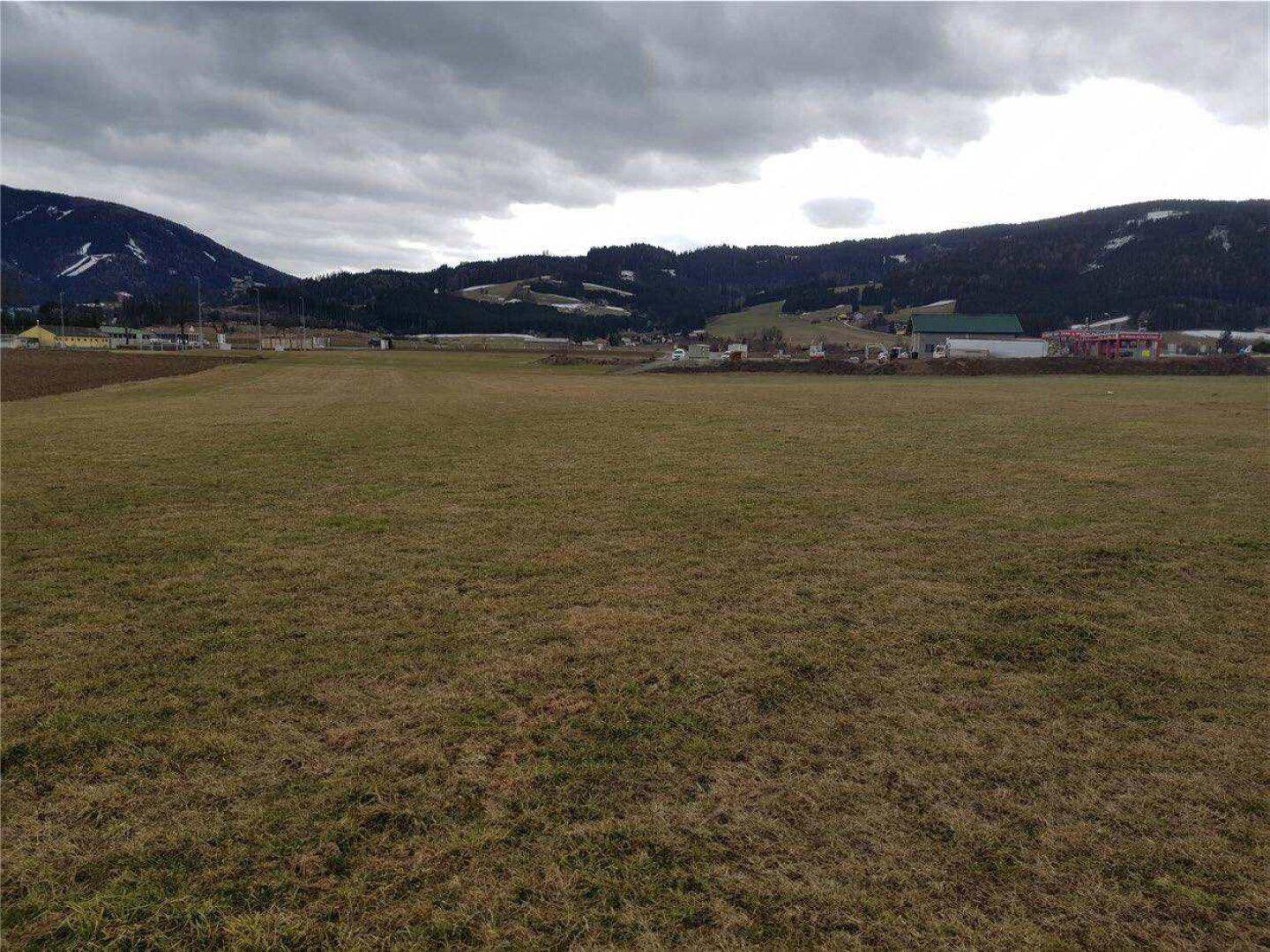 Weitläufiges, unbebautes Feld mit Blick auf bewaldete Hügel und schneebedeckte Berge im Hintergrund unter bewölktem Himmel.