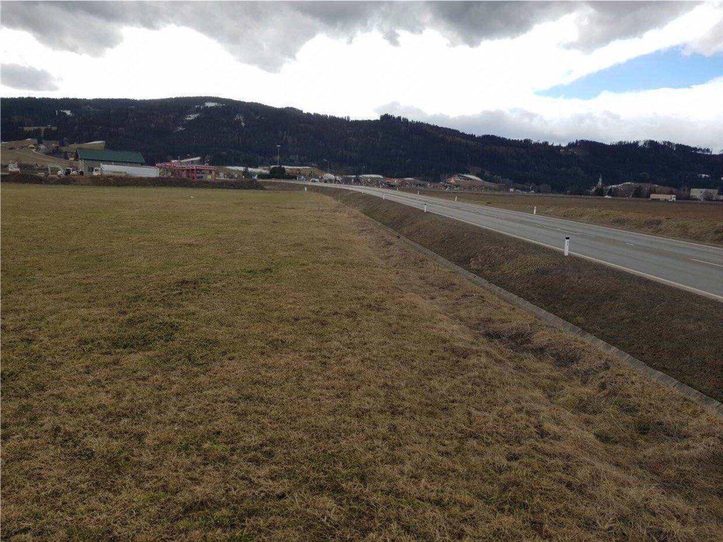 Unbebautes Feld entlang einer Landstraße mit Blick auf die umliegende ländliche Landschaft und Berge am Horizont.