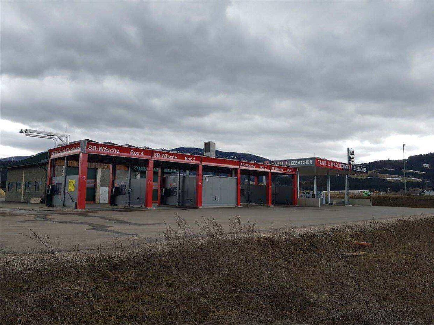 Gebäude einer Tankstelle und Waschanlage mit roten Akzenten und einem großen Parkplatz unter einem bewölkten Himmel.
