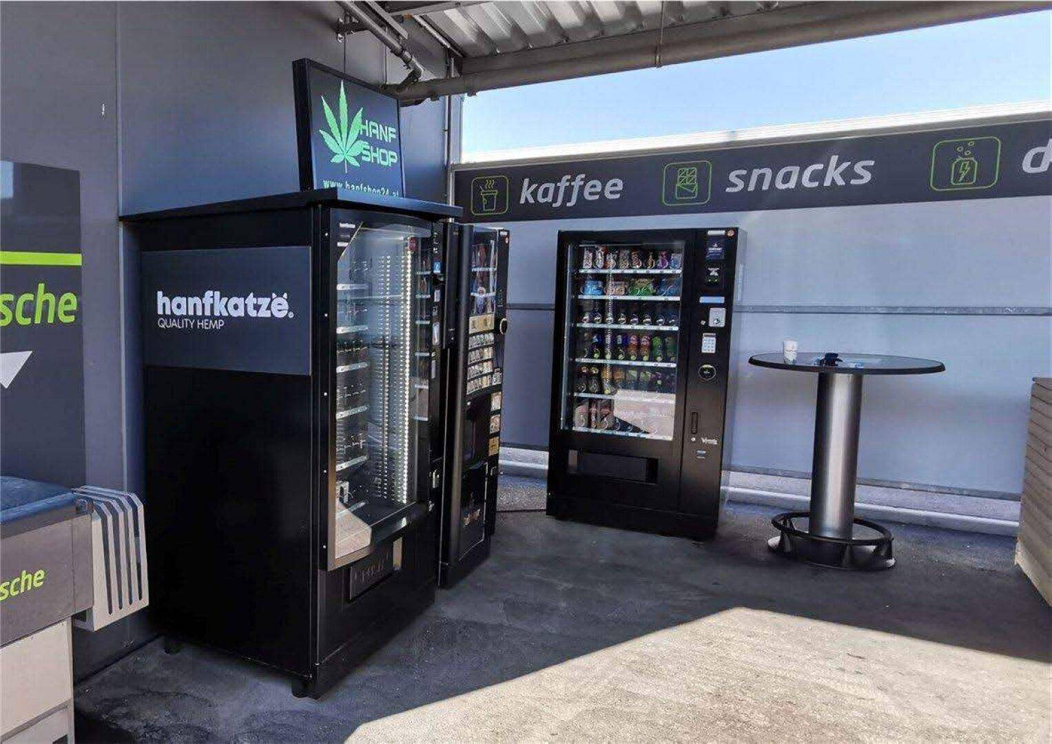 Überdachter Bereich mit Verkaufsautomaten für Kaffee, Snacks und Hanfprodukte sowie einem Stehtisch.