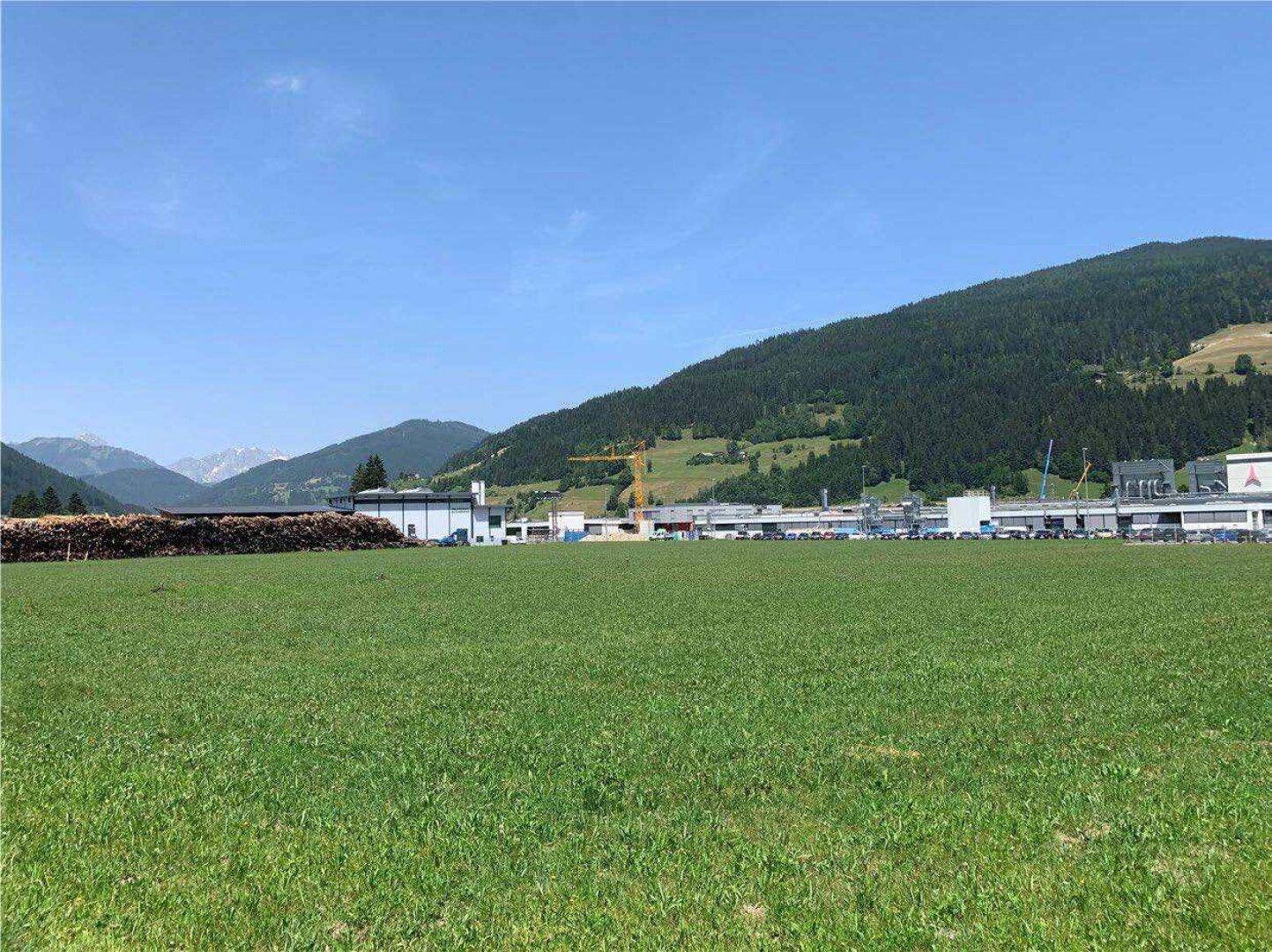 Weitläufiges grünes Feld mit Industriegebäuden und Bergen im Hintergrund unter einem klaren blauen Himmel.
