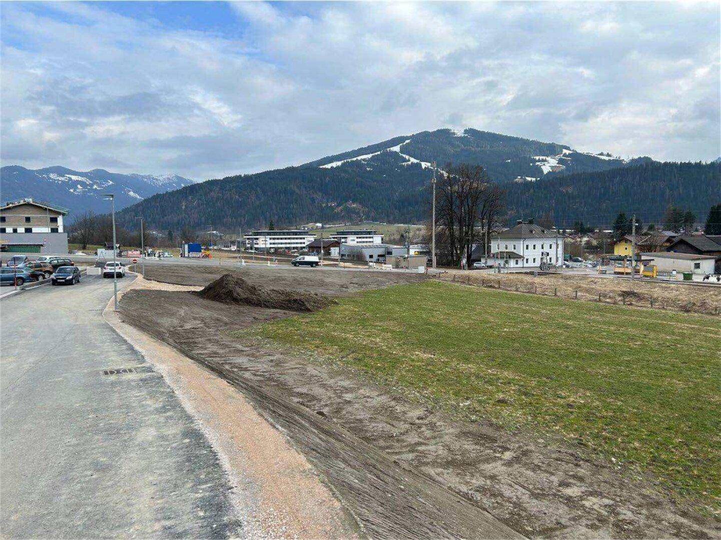 Ungenutztes Grundstück mit einer Straße im Vordergrund und schneebedeckten Bergen im Hintergrund unter bewölktem Himmel.