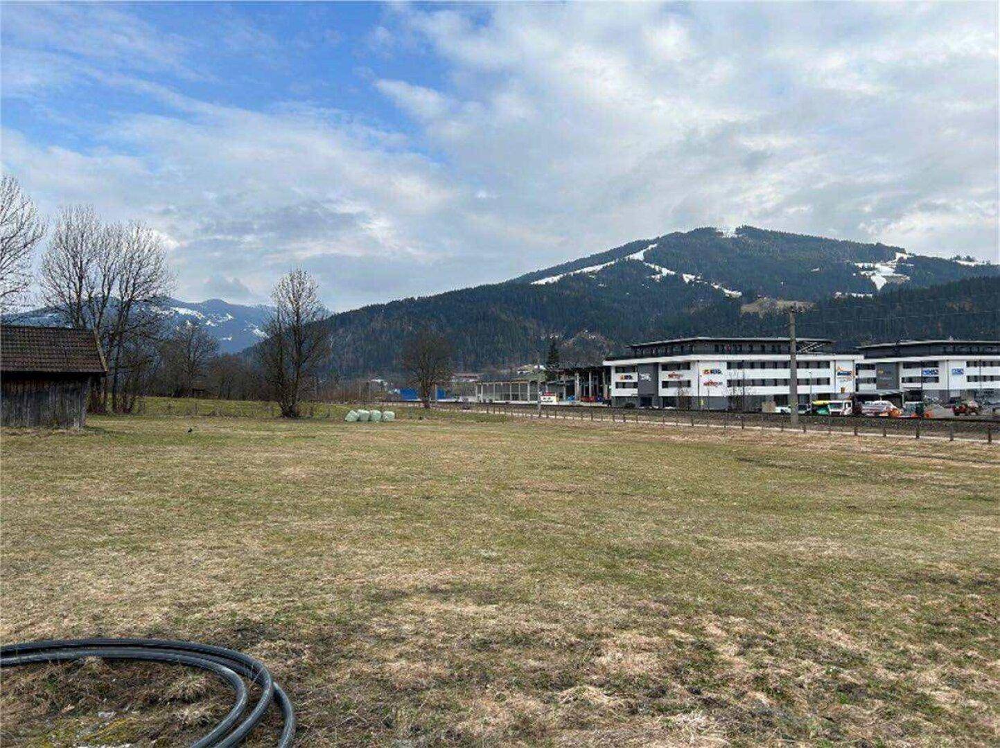 Weitläufiges grünes Feld mit Gebäuden und schneebedeckten Bergen in der Ferne unter einem bewölkten Himmel.