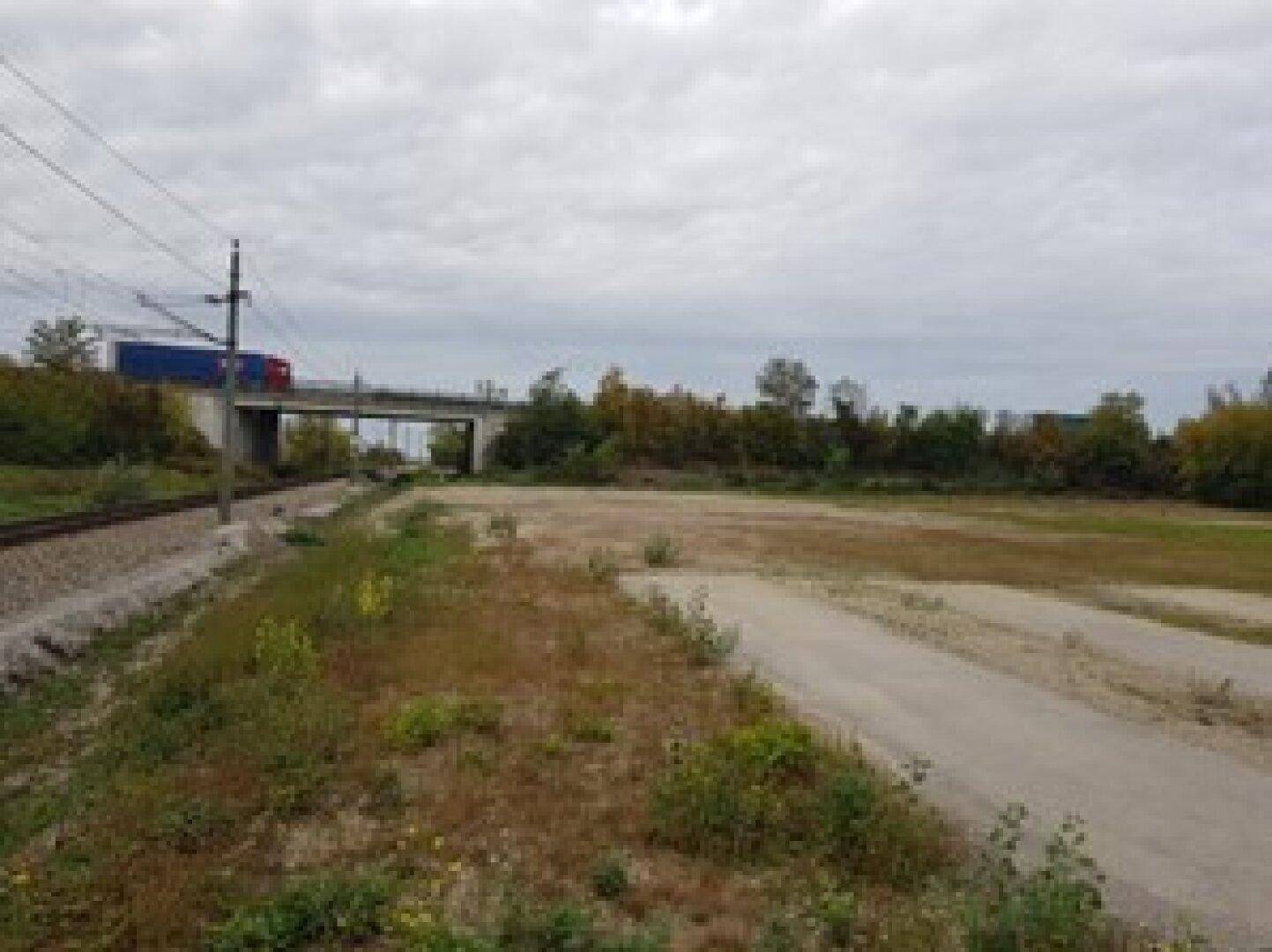 Unbebautes Grundstück mit Blick auf eine Eisenbahnlinie und eine Autobahnbrücke im Hintergrund unter bewölktem Himmel.