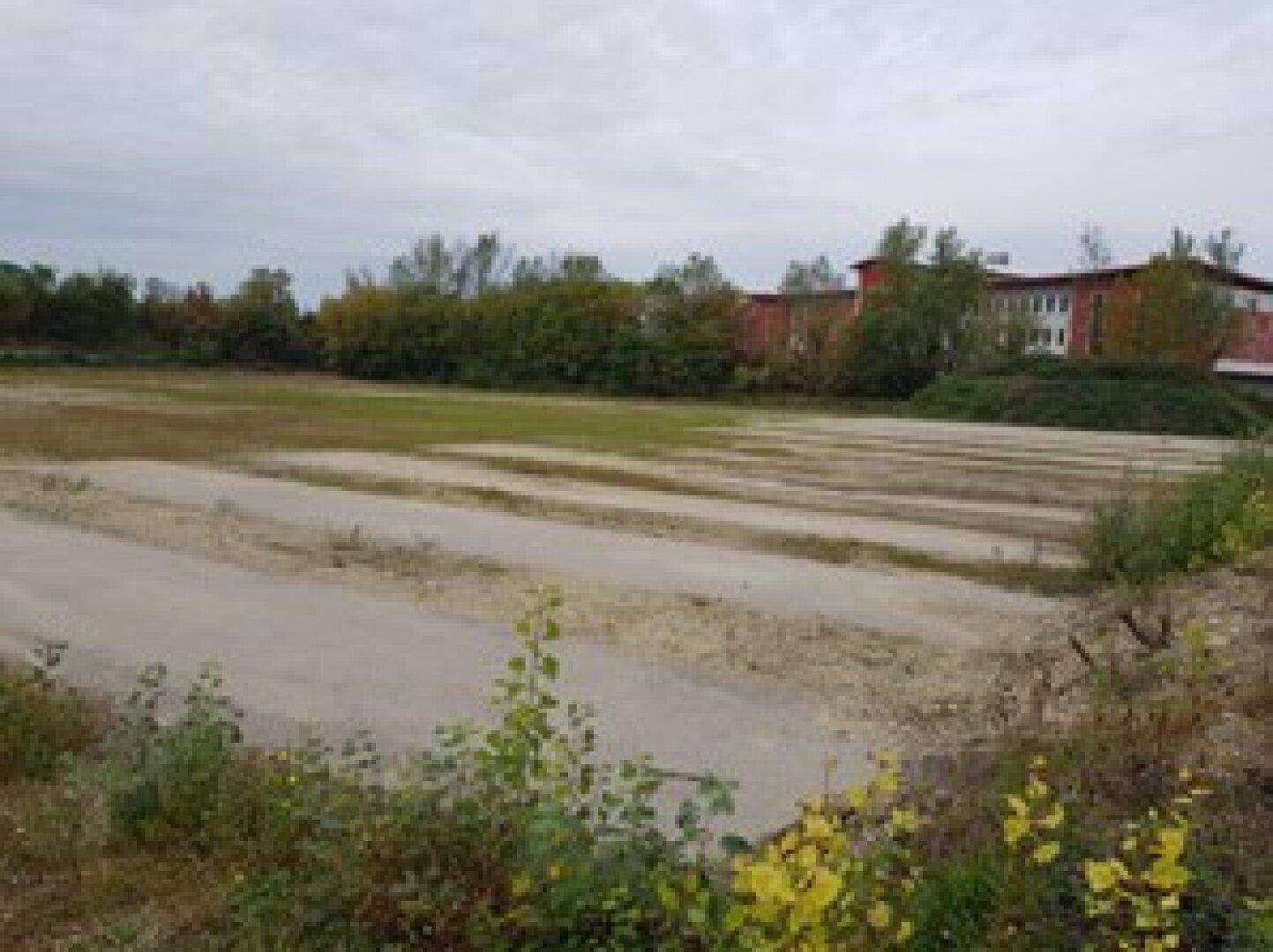 Weite Ansicht des unbebauten Grundstücks mit spärlicher Bepflanzung und Bäumen im Hintergrund unter bewölktem Himmel.