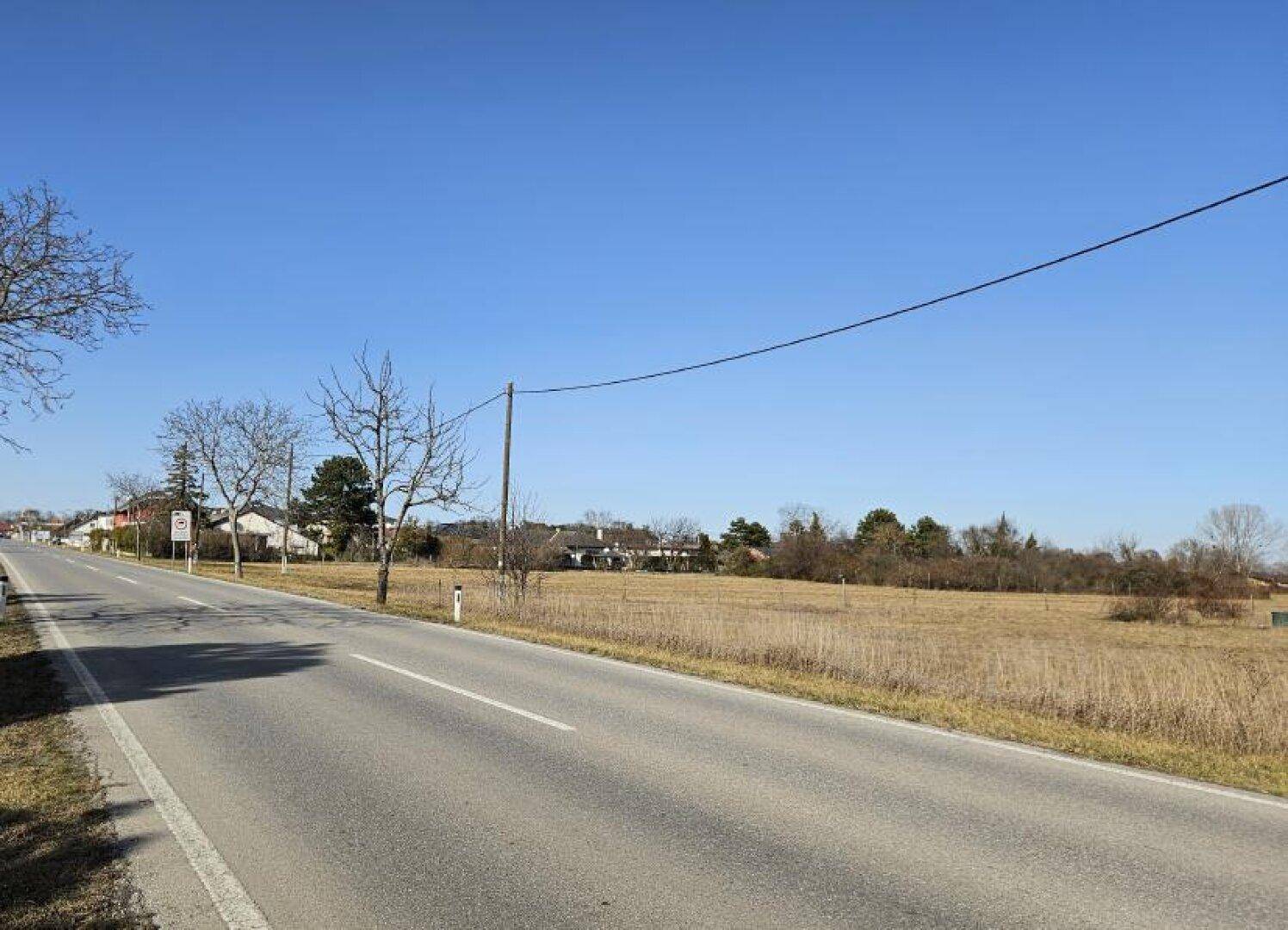 Blick auf eine Landstraße mit angrenzendem Feld und Häusern in der Ferne unter blauem Himmel.