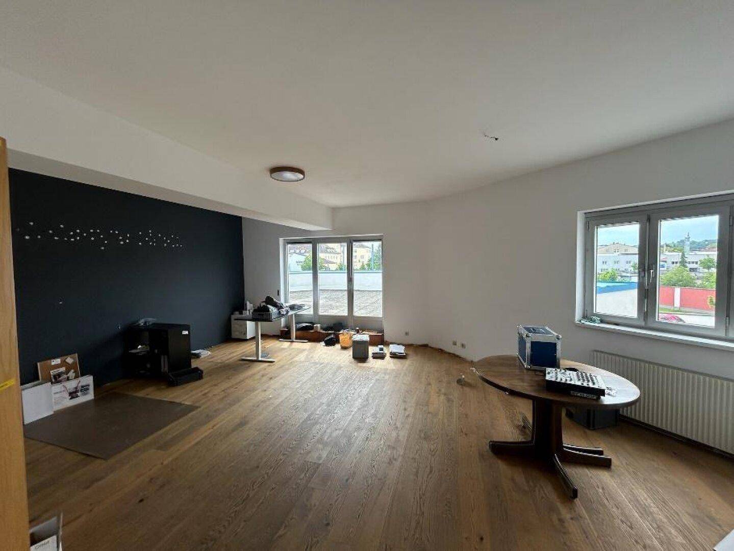 Geräumiger Raum mit Holzboden, einer dunklen Akzentwand und mehreren Fenstern für viel Tageslicht.