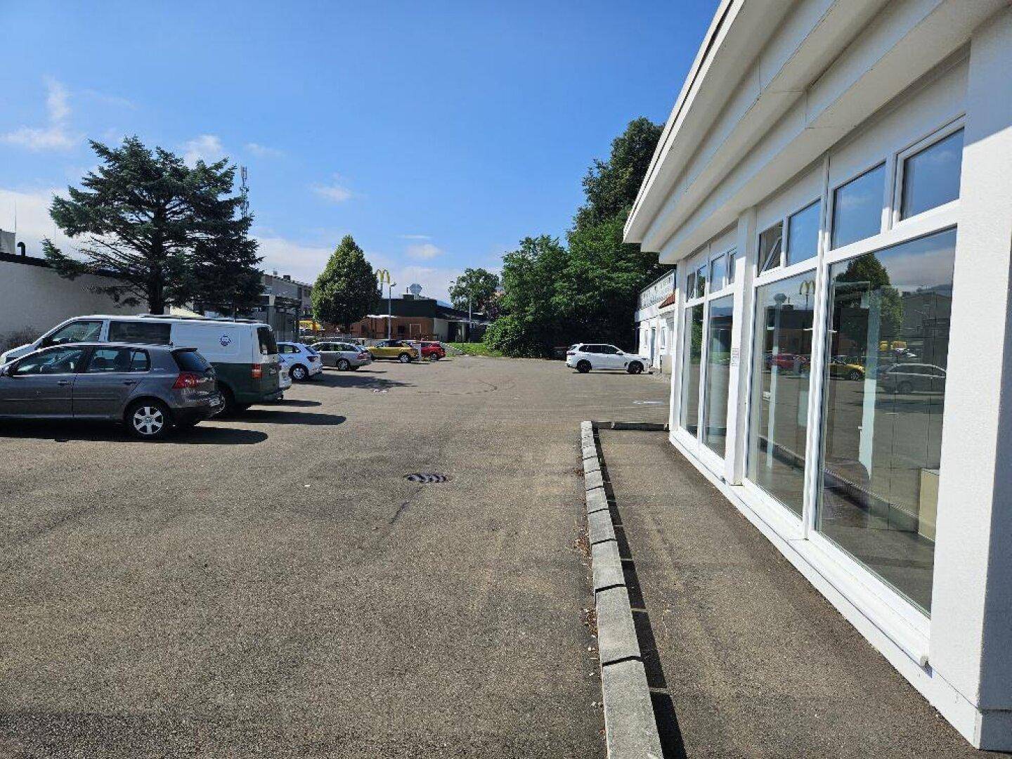 Ansicht des Parkplatzes neben dem Geschäftsgebäude mit großen Schaufenstern und Bäumen im Hintergrund.