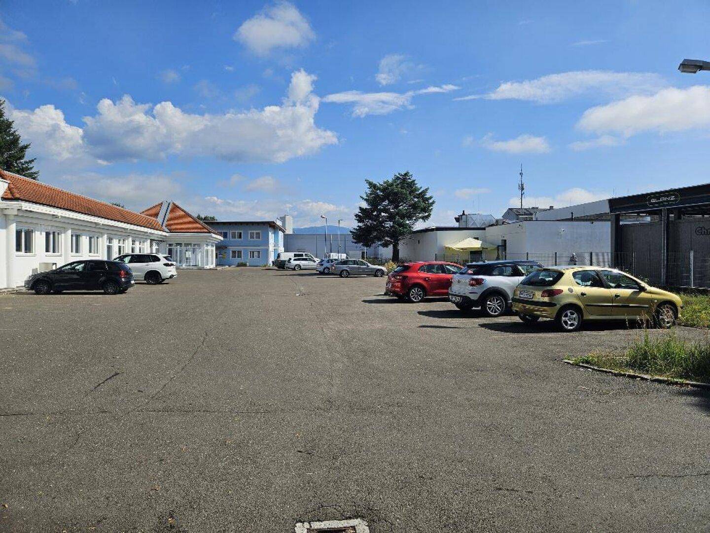 Großer asphaltierter Parkplatz mit mehreren geparkten Autos und umliegenden Gebäuden unter blauem Himmel.