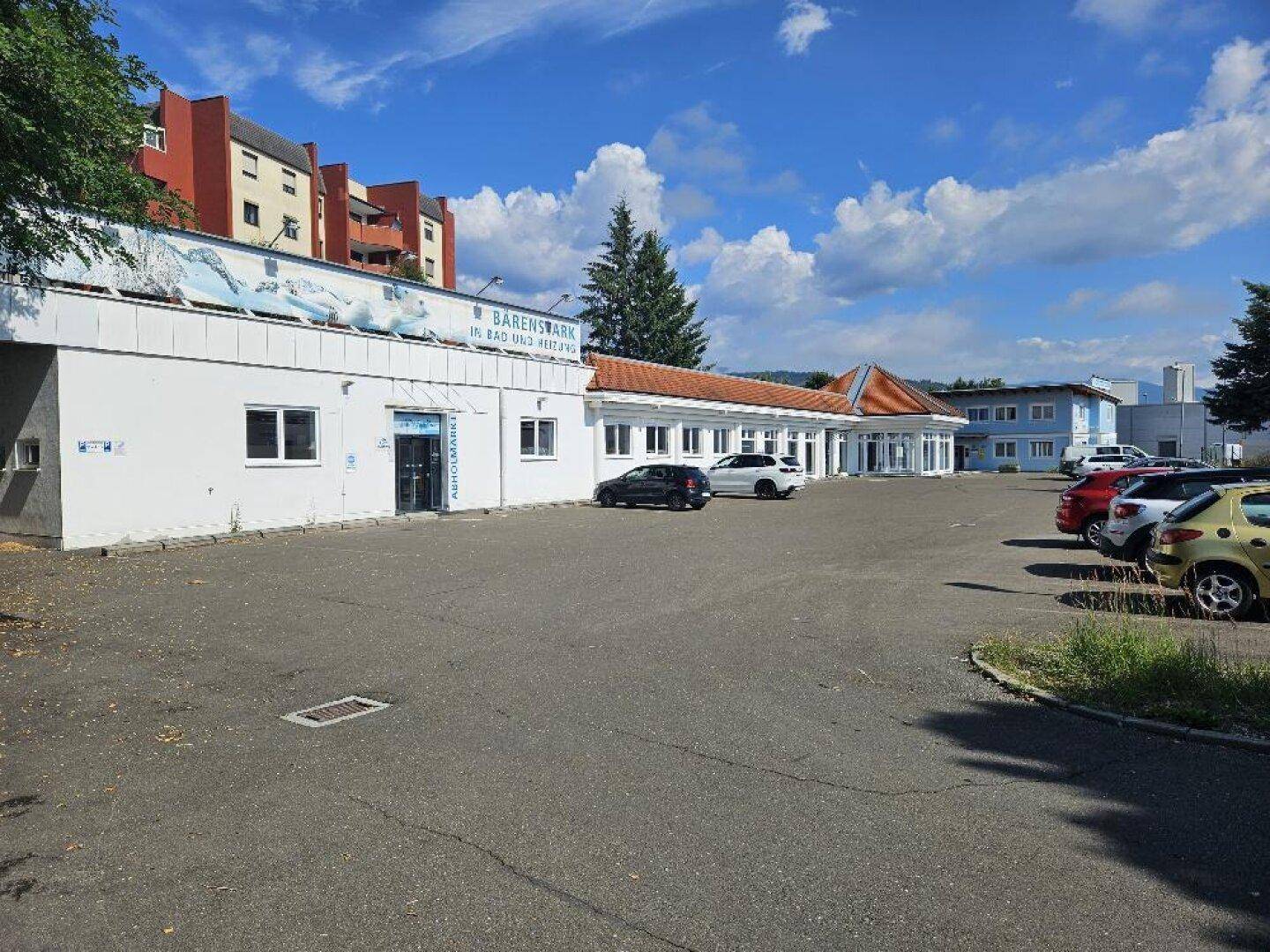 Weitläufiger Parkplatz vor einem Geschäftsgebäude mit mehreren Eingängen und einem blauen Himmel.