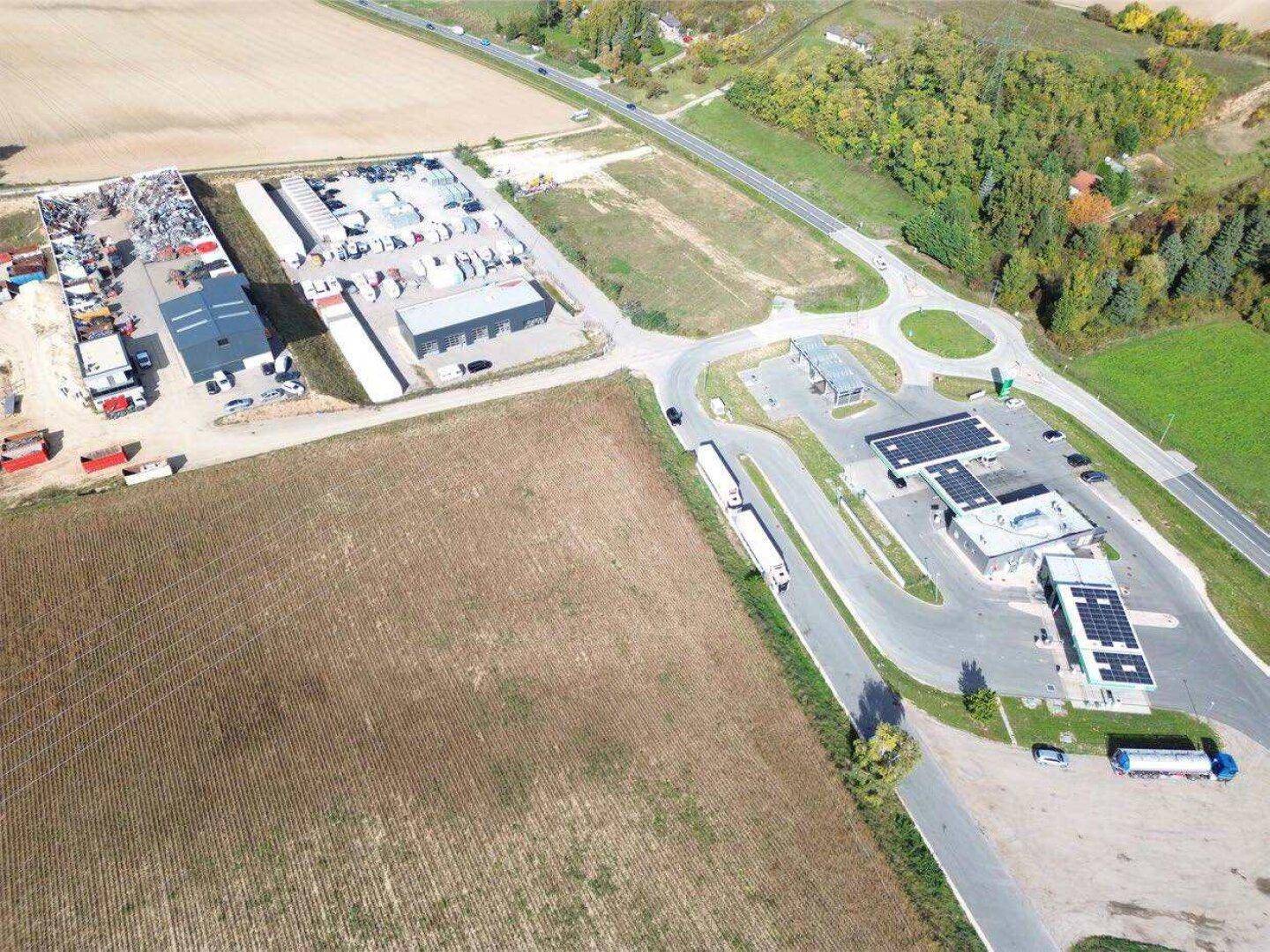 Luftbild des Gewerbegebiets mit Tankstelle, Parkplätzen und angrenzenden Feldern unter klarem Himmel.