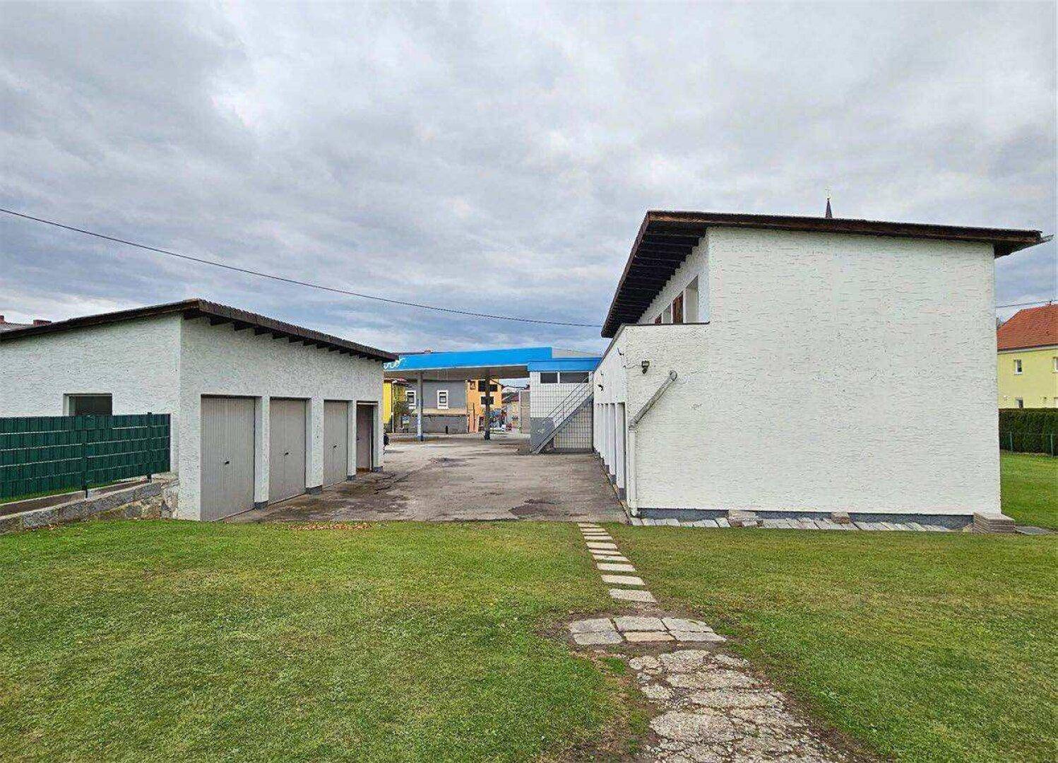 Rückansicht des Anwesens mit zwei weißen Gebäuden, Garagen und einem gepflegten Rasen mit Steinweg.