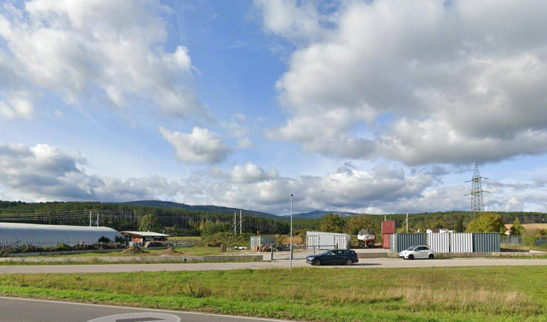 Weitläufiges Außengelände mit Lagercontainern, Straße und bewaldeten Hügeln unter blauem Himmel.