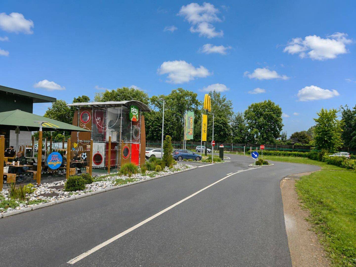 Außenbereich des McDonald's mit Terrasse, Spielplatz und Drive-in-Spur unter blauem Himmel.