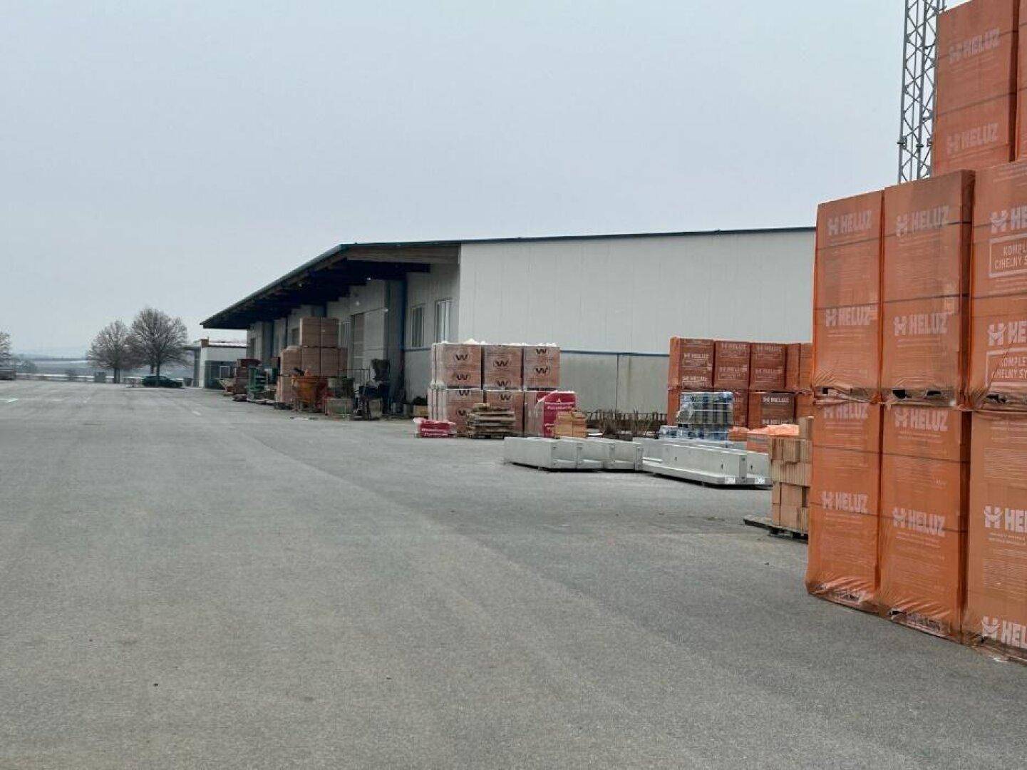 Außenansicht einer Lagerhalle mit großem gepflasterten Hof und gestapelten Baumaterialien unter bewölktem Himmel.