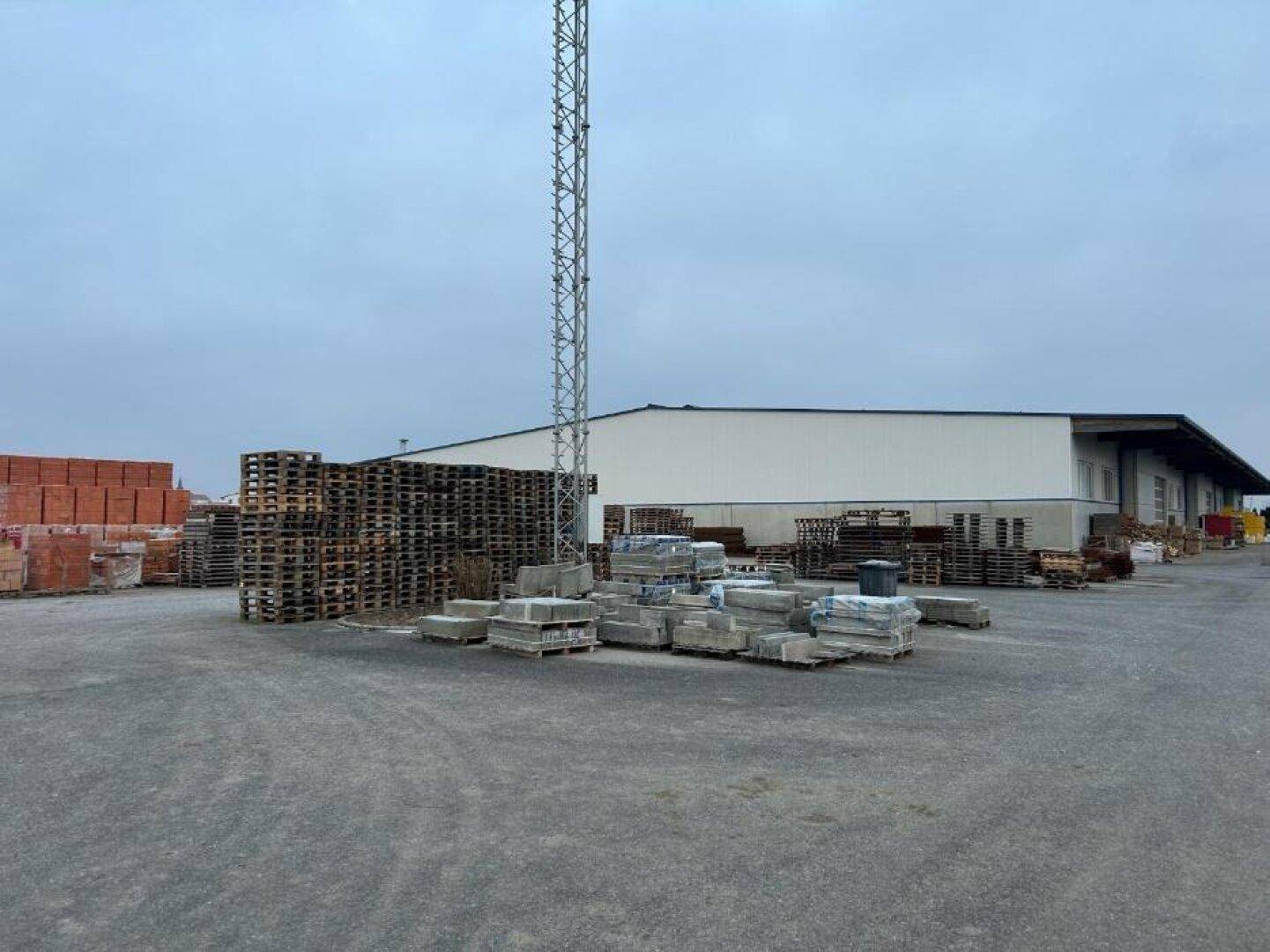 Außenbereich der Lagerhalle mit großen Stapeln von Paletten und Baumaterialien unter bewölktem Himmel und einem Mast.