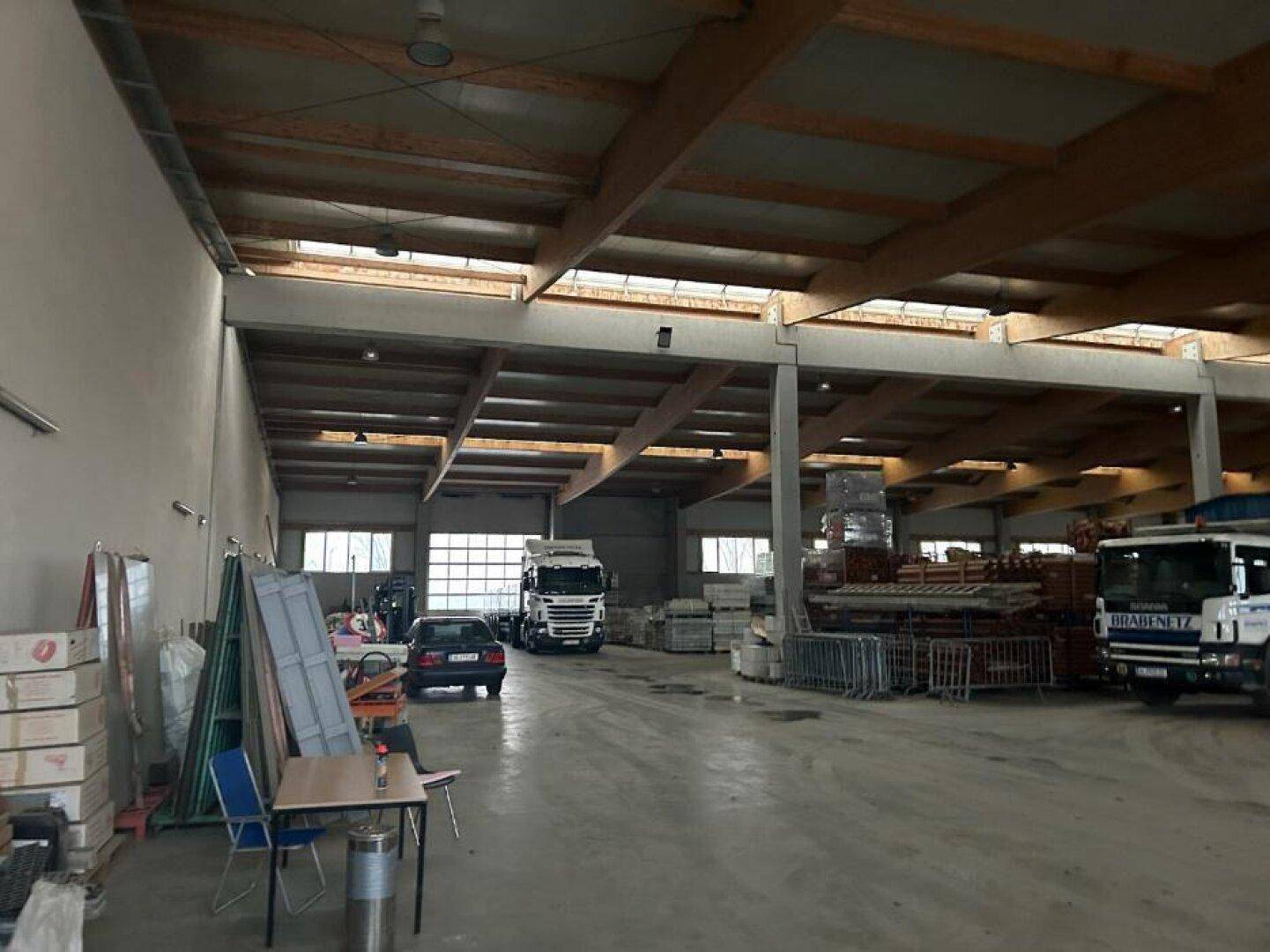Innenansicht der Lagerhalle mit hohen Decken, großen Toren und Fenstern, sowie abgestellten Fahrzeugen und Materialien.