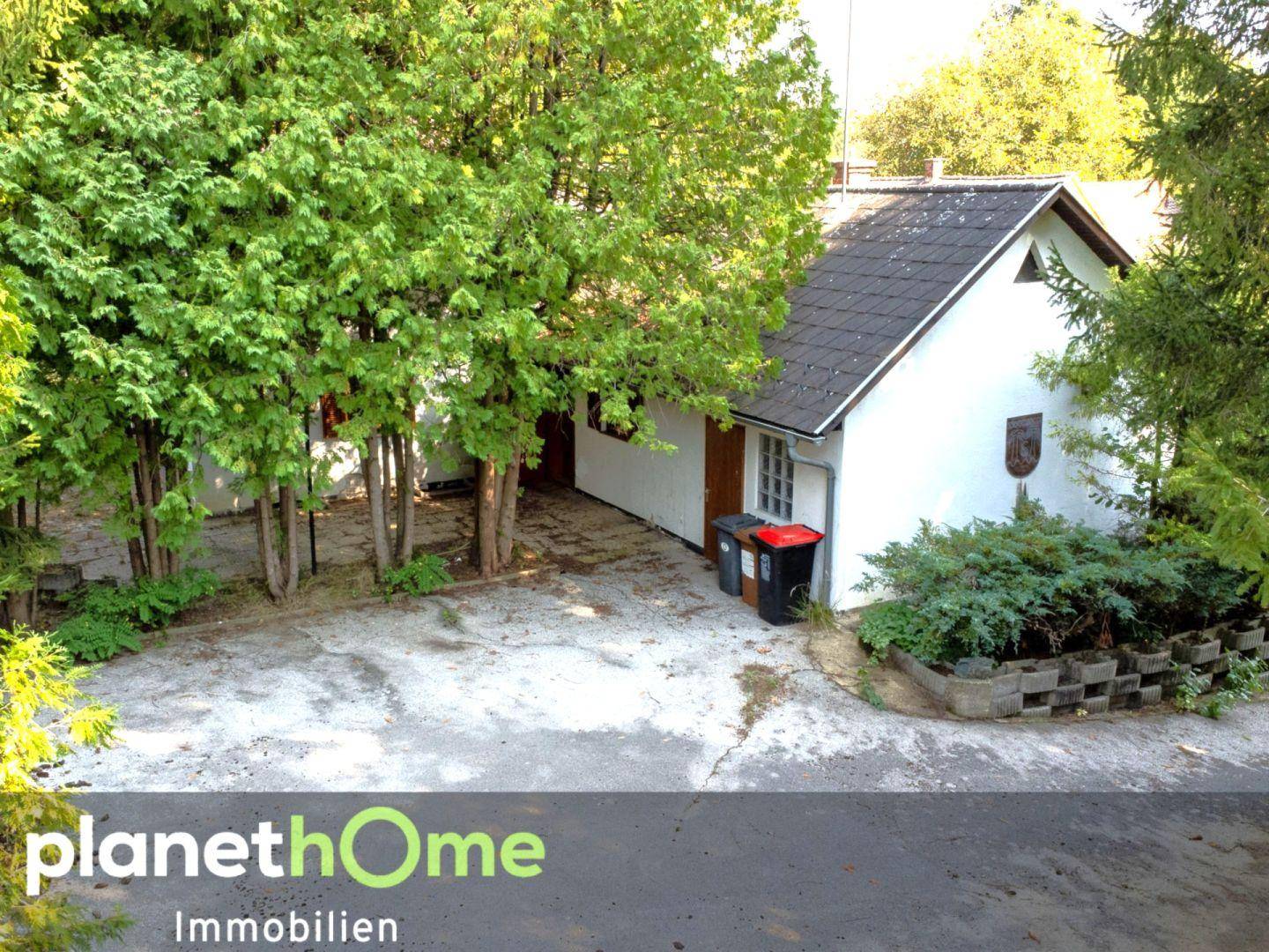 Außenansicht eines kleinen weißen Hauses mit dunklem Dach, umgeben von Bäumen und einer gepflasterten Fläche.