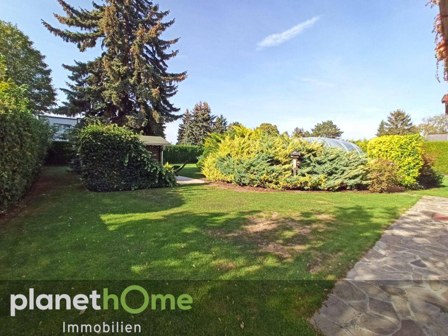 Gepflegter Garten mit Rasen, Sträuchern und einem kleinen Gartenhaus unter blauem Himmel.