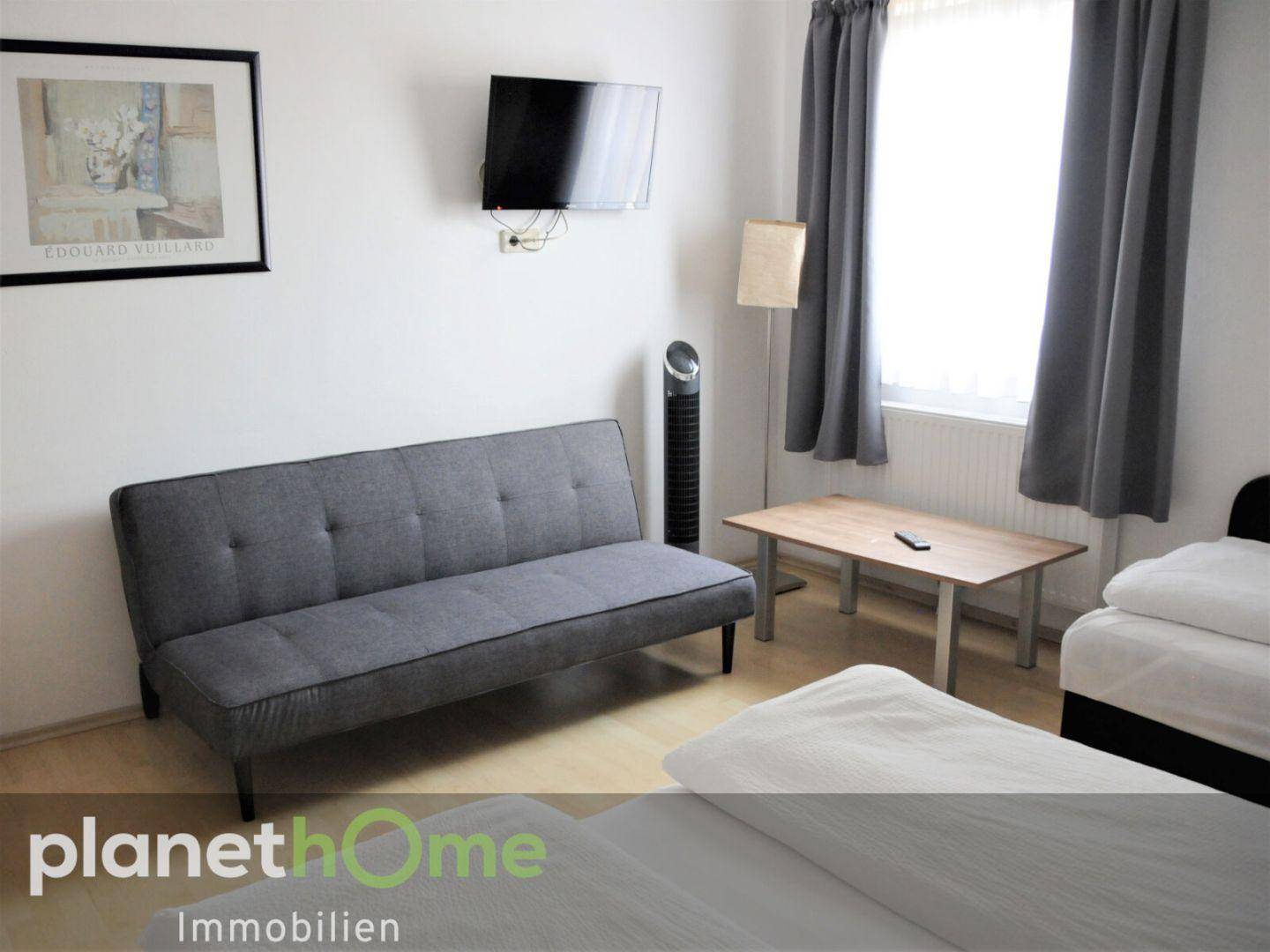 Helles Schlafzimmer mit grauem Sofa, Fernseher und einem Bett, einfach eingerichtet.